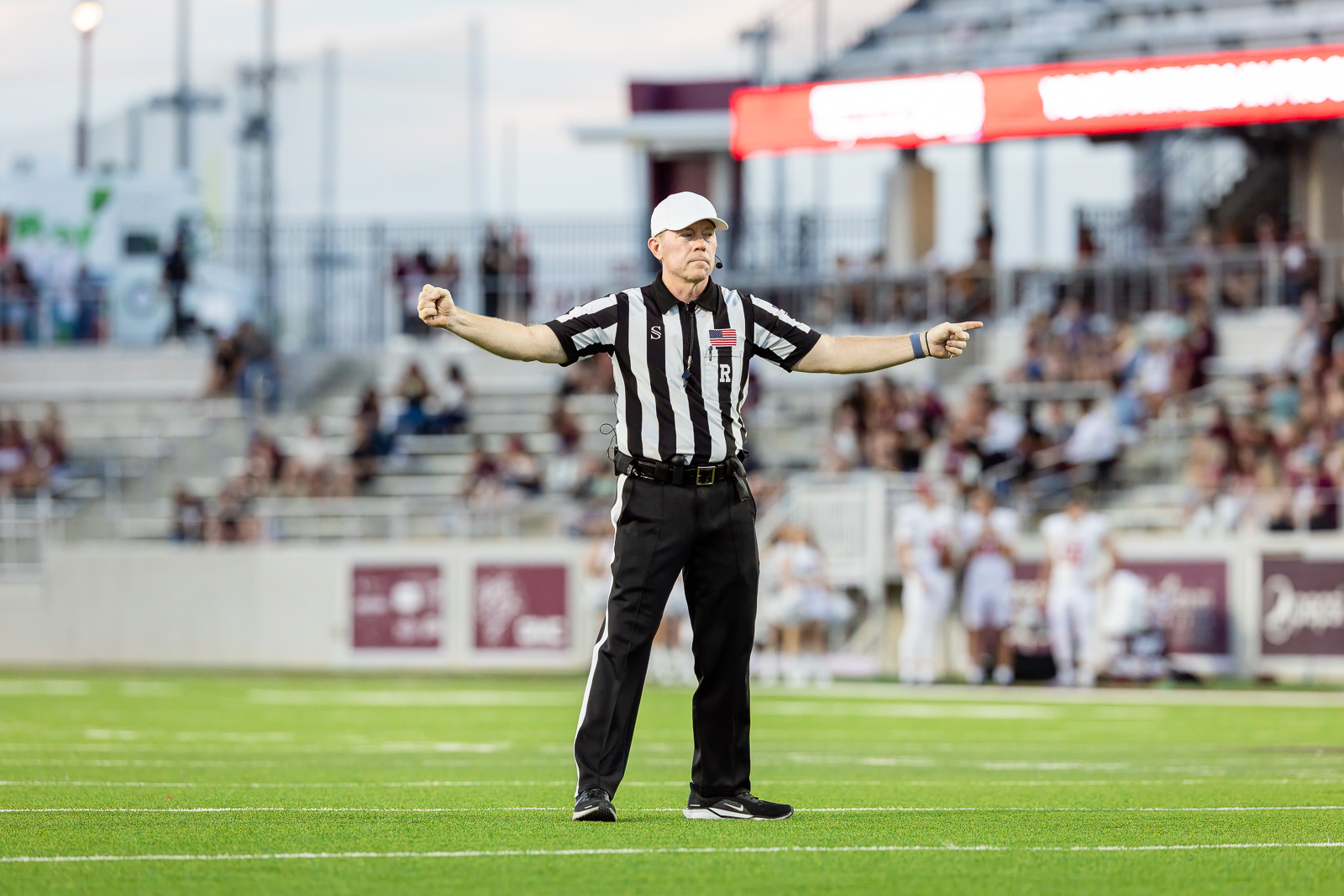 western-colorado-vs-wtamu-jpegJoe-pps-57 | Press Pass Sports Football referee on a green field signals a call with arms outstretched, crowd blurred in the stands behind.