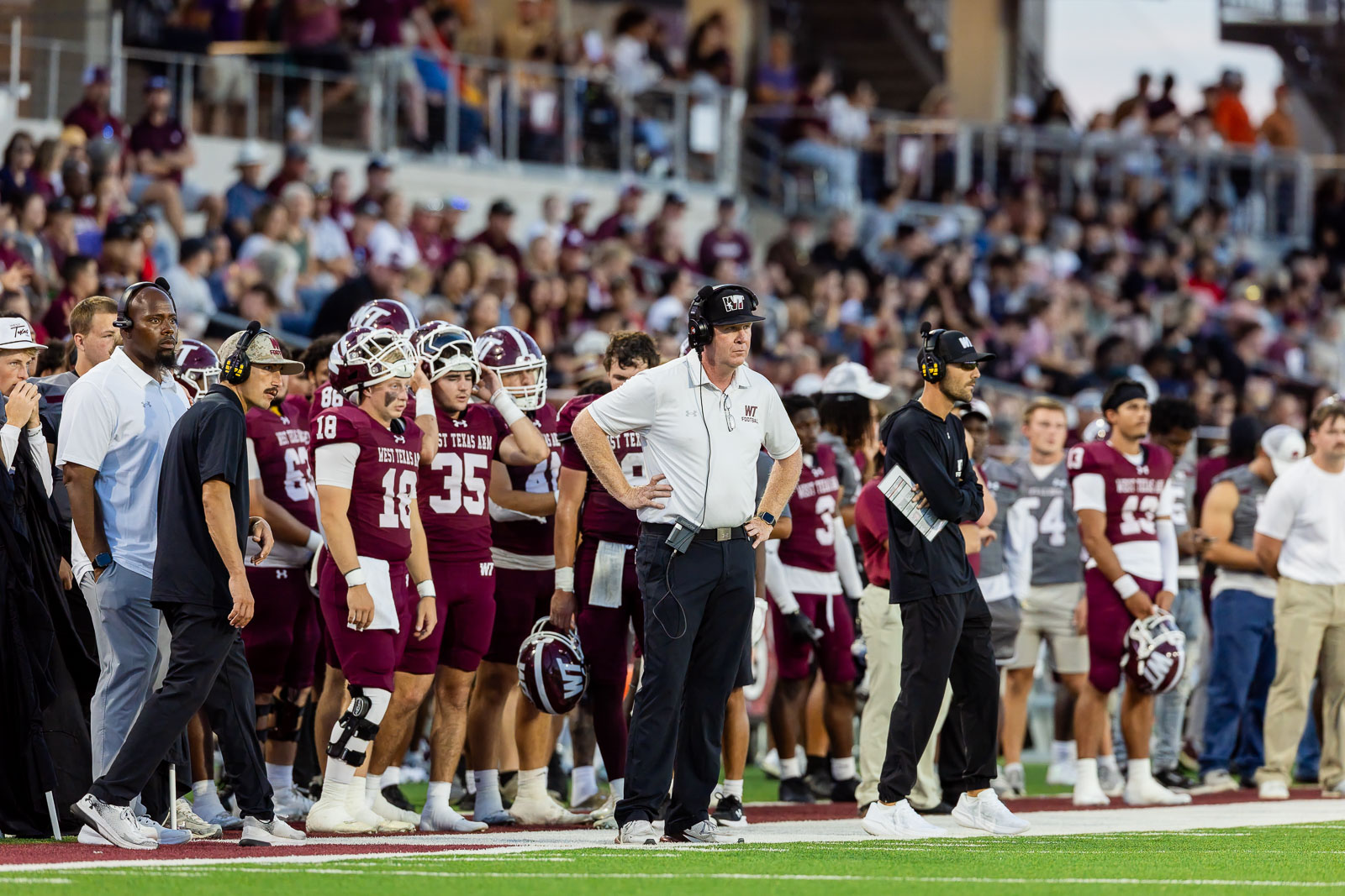 western-colorado-vs-wtamu-jpegJoe-pps-58 | Press Pass Sports Football team on the sideline during a game, coach wearing a headset and white polo amid maroon uniforms and a crowded stadium behind.