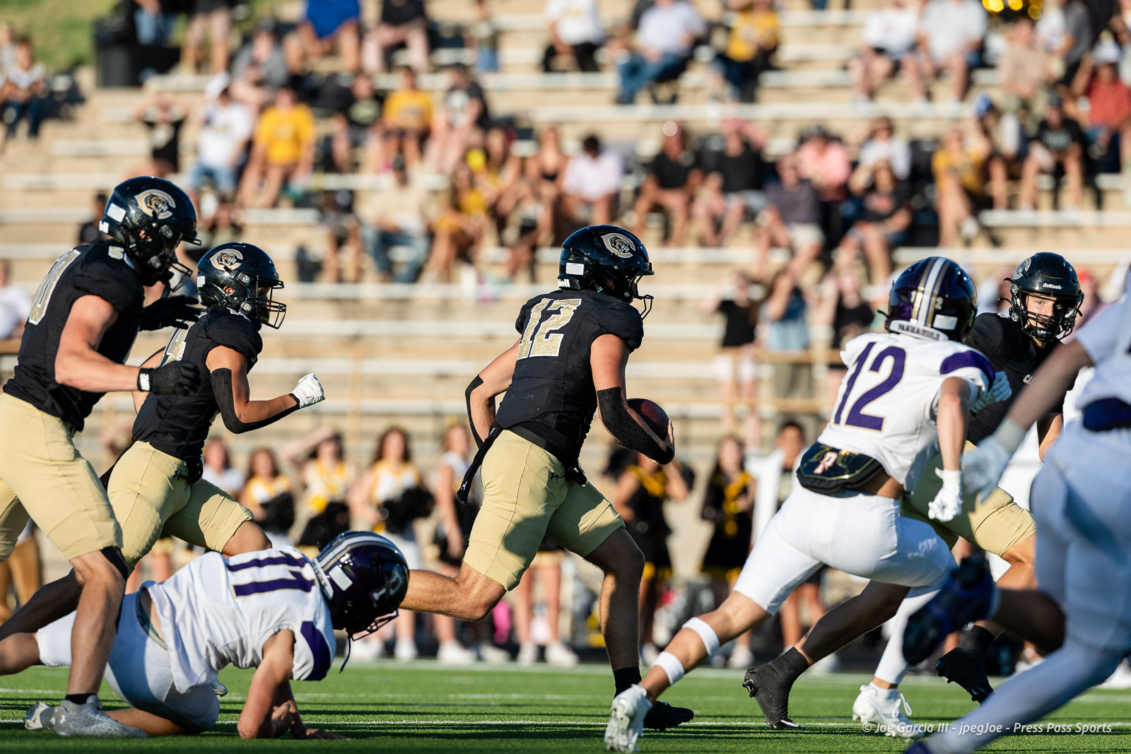 panhnandle-vs-canadian-football-jpegJoe-pps-2 | Press Pass Sports Football game action: a player in black jersey U12 runs with the ball while defenders close in on a sunny sideline stadium.