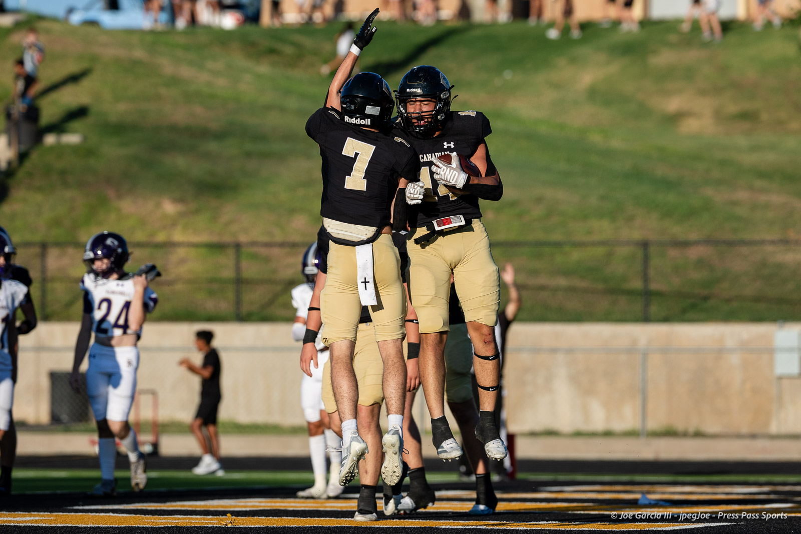 panhnandle-vs-canadian-football-jpegJoe-pps-5 | Press Pass Sports Two football players in black and gold jerseys leap and high-five in midair on a football field as teammates celebrate a score, with others and a grassy hill in the background.