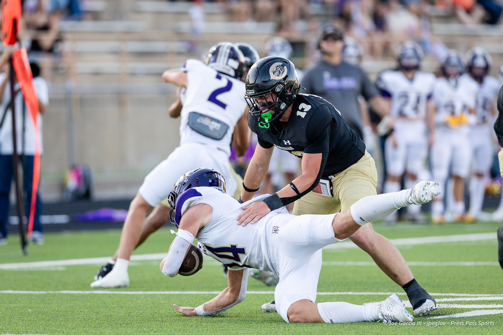 panhnandle-vs-canadian-football-jpegJoe-pps-8 | Press Pass Sports Football players collide on a turf field as a defender in black and gold tackles a ball carrier in white and purple.