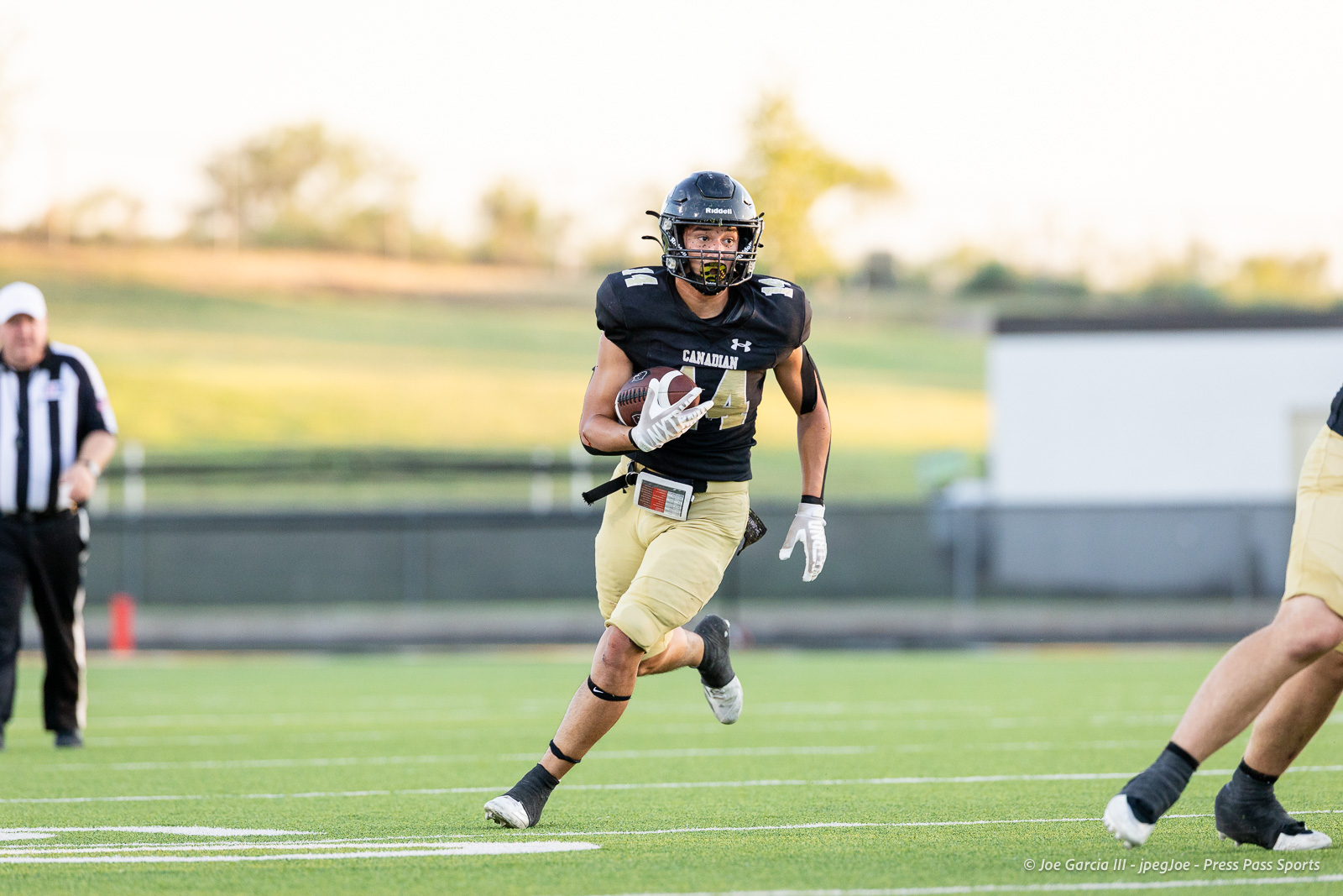panhnandle-vs-canadian-football-jpegJoe-pps-13 | Press Pass Sports Football player in black jersey sprinting with the ball on a green field, referee in the background.