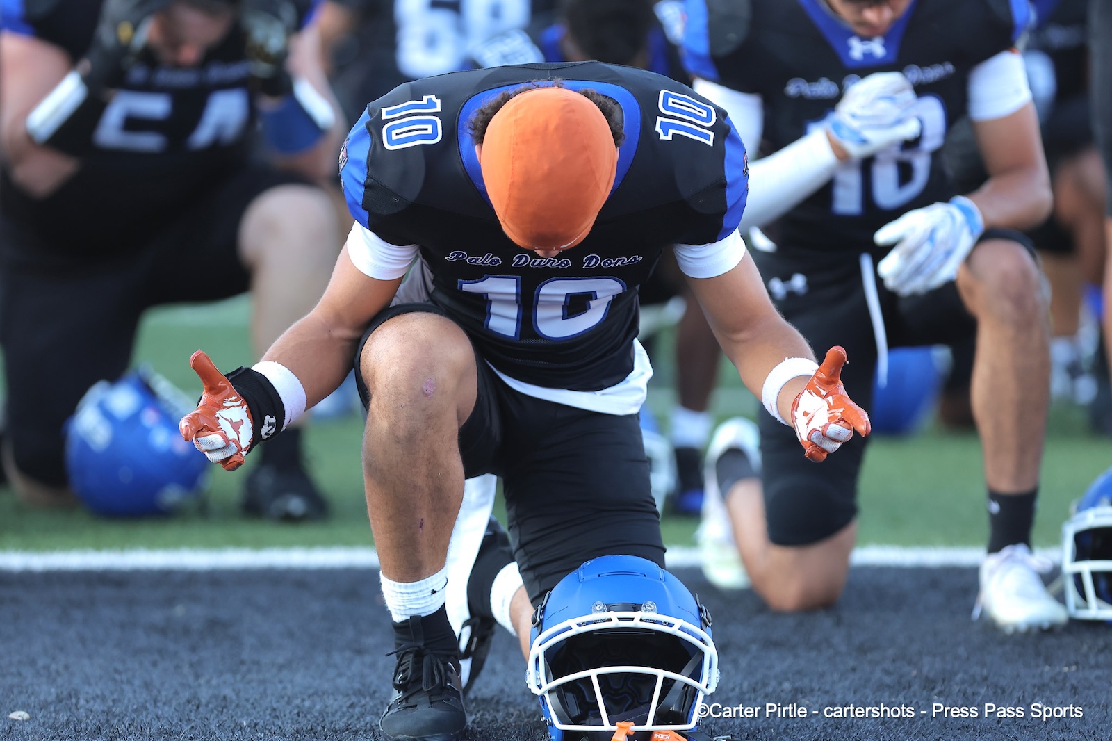 Football player in black/blue uniform kneels on a turf field, head bowed over a fallen blue helmet with an orange helmet nearby.