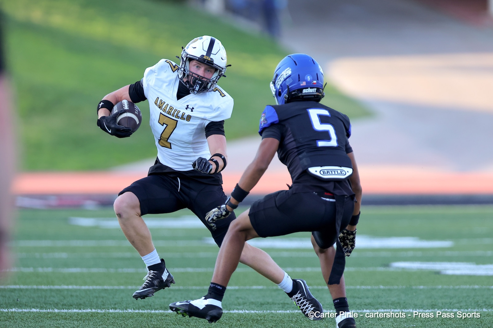 Football player in white number 7 with ball rushing past a blue-jersey defender (#5) on a grassy field, during a game, moment of action.