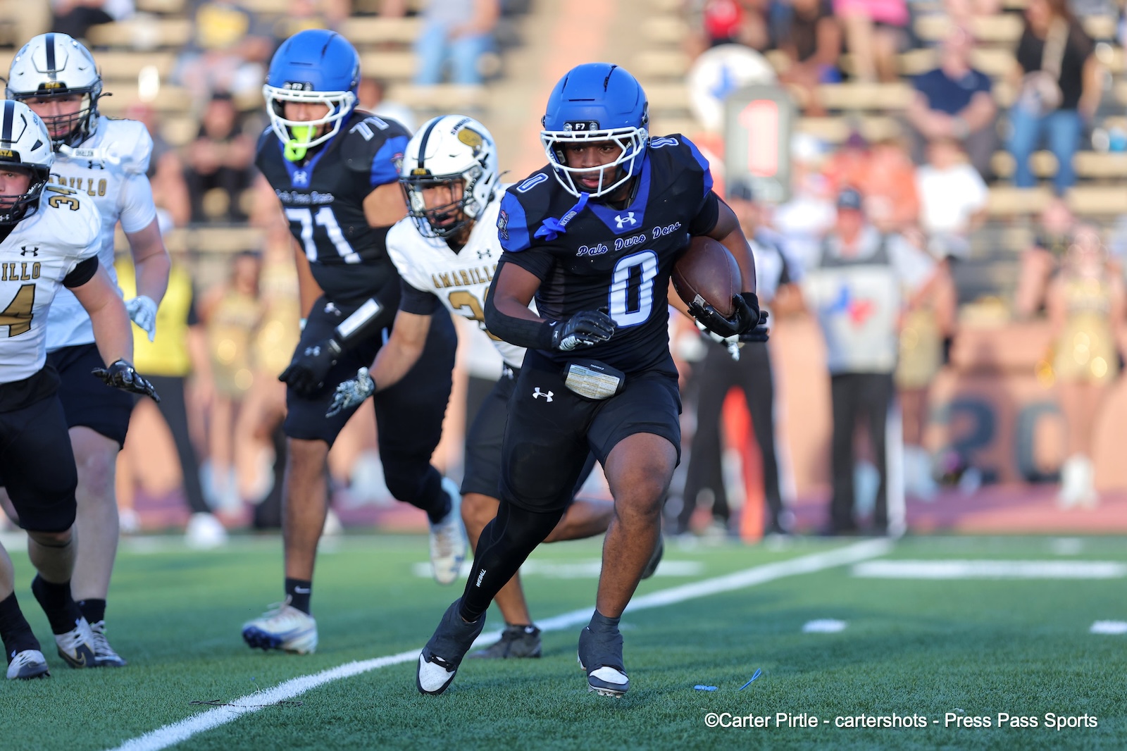 Athlete in blue uniform carrying a football while running down the field, pursued by opponents.