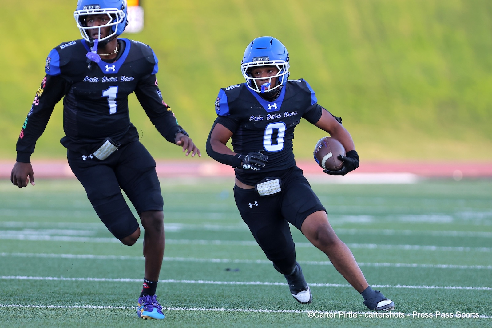Two football players in black and blue uniforms sprint on a turf field; the player wearing number 0 holds the ball and looks ahead while his teammate runs beside him.