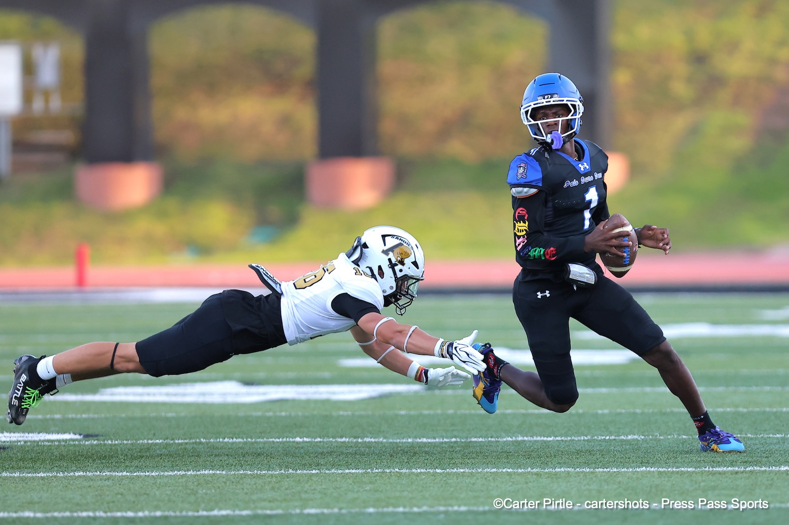 Football player in black uniform holds ball while running, pursued by a diving defender in white and black on a green field.