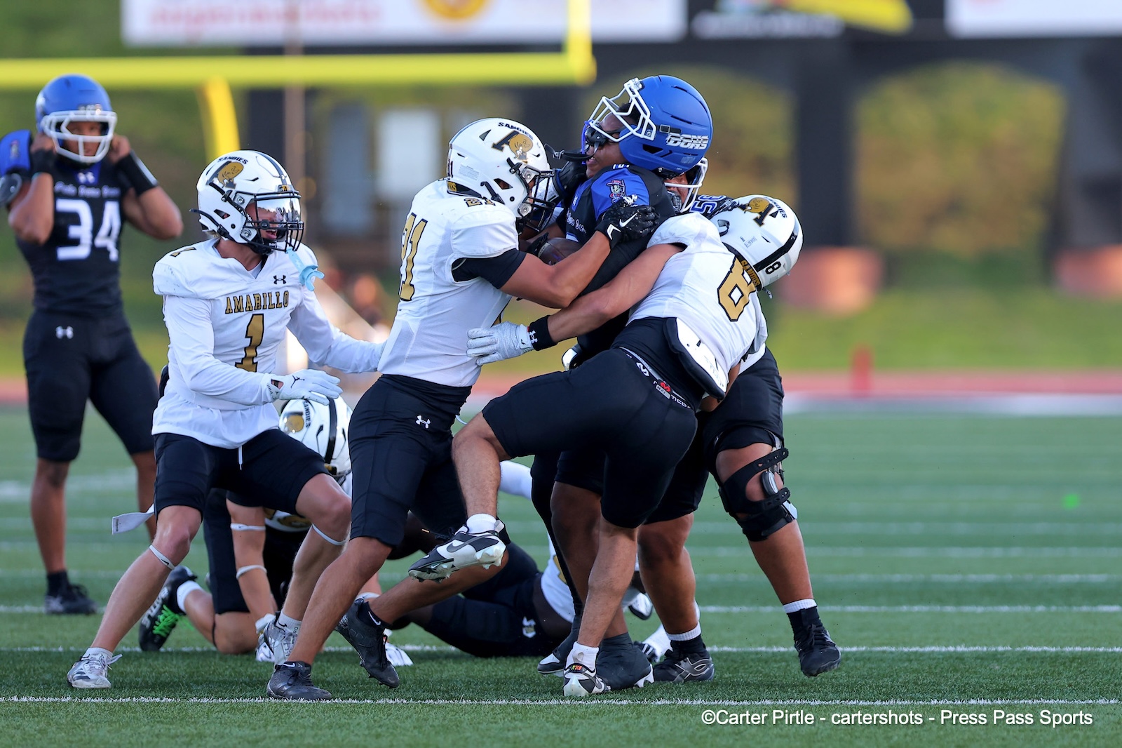 Football players in white and black tackle a ball carrier in blue helmet on a turf field during a game.