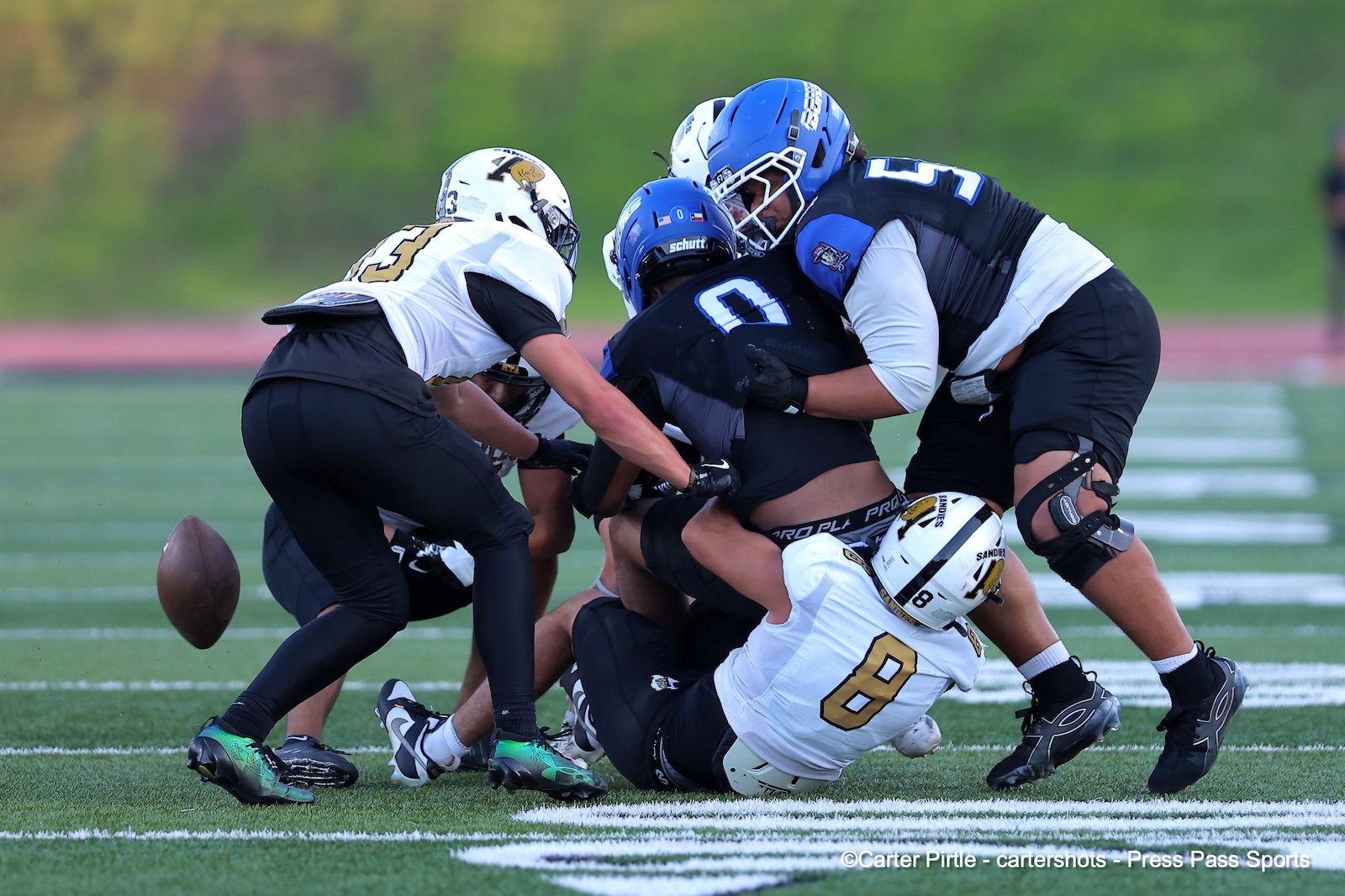 Football players tackle for the ball in a crowded scrum on a green turf field, ball loose nearby.
