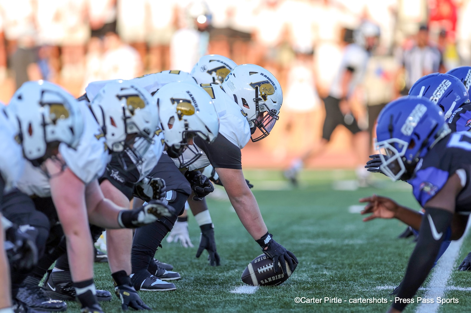 Football line of scrimmage: center snapping the ball to the quarterback while white-helmeted linemen block against a blue defense.