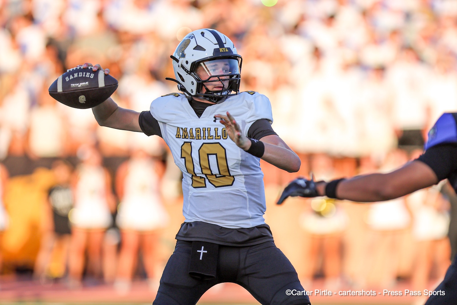 Quarterback wearing a white jersey with number 10 and a white helmet throws a pass during a football game, with a blurred crowd in the background.