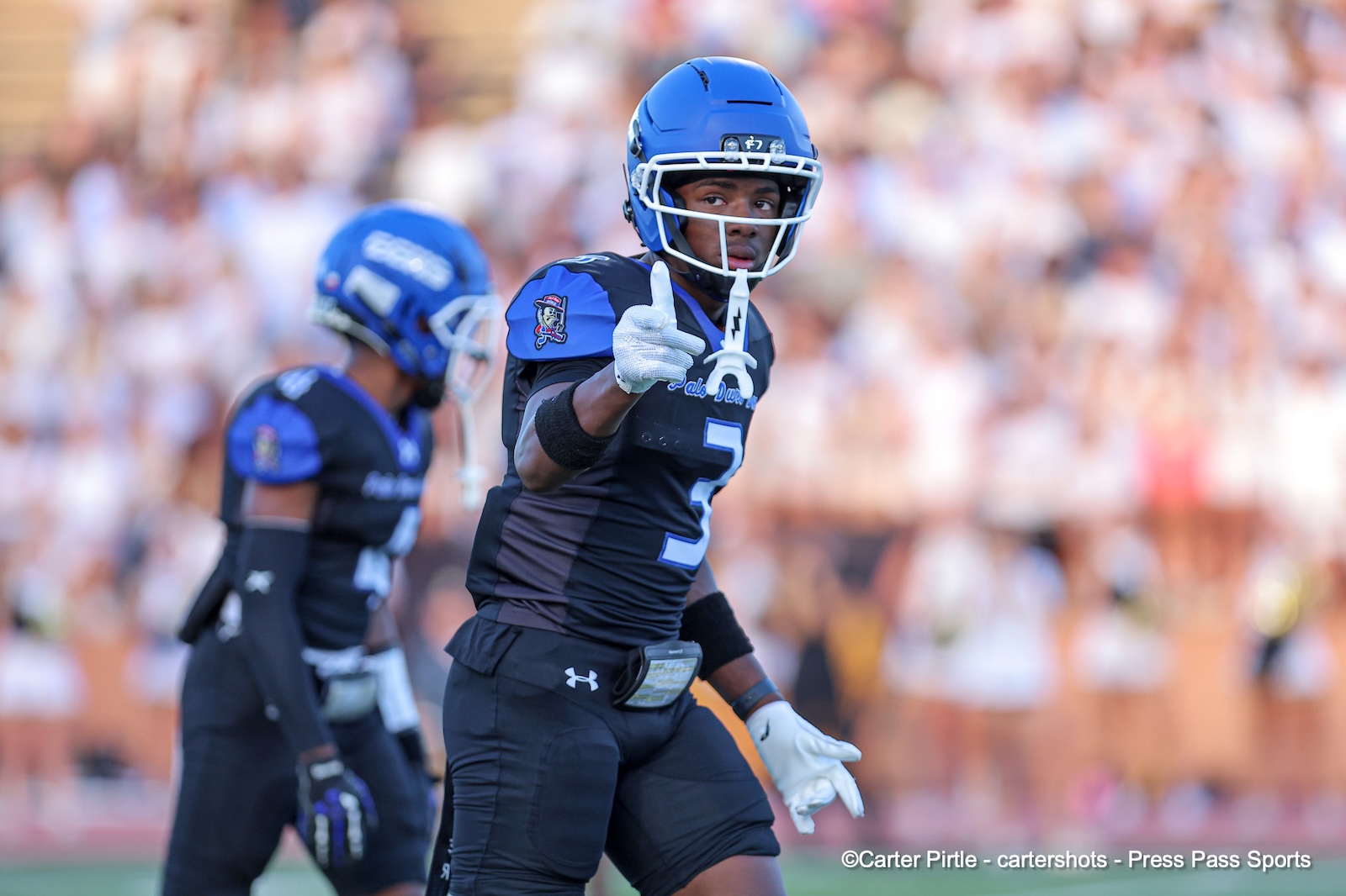 Football player in blue helmet and jersey #3 gives a thumbs-up on the field, with a blurred stadium crowd behind him.