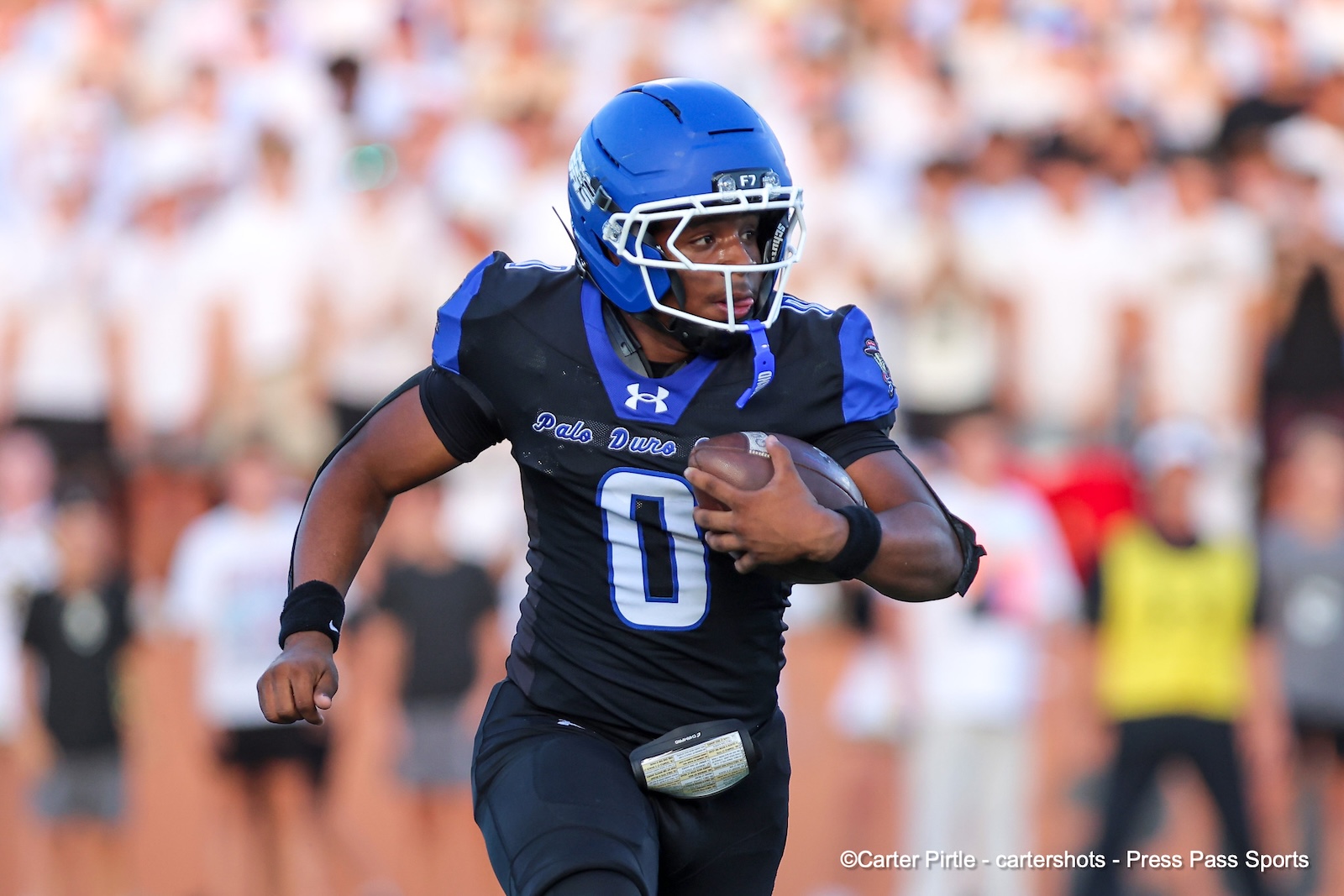 Football player in a blue helmet and black-blue jersey running with ball during a game, crowd blurred in the background, number 0 visible on jersey.