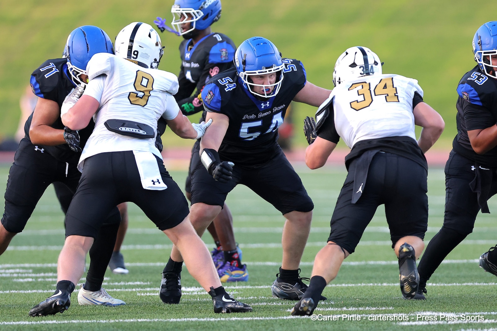 Football players in blue and white uniforms collide at the line of scrimmage on a green field.