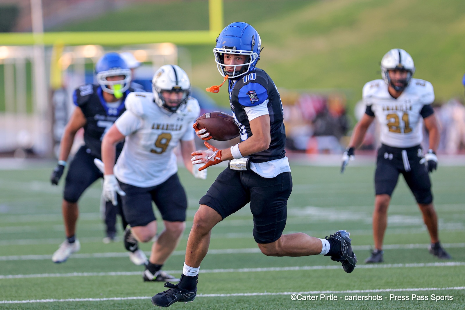 Male football player in a blue helmet carries the ball while running on a green turf field, chased by defenders in white jerseys.