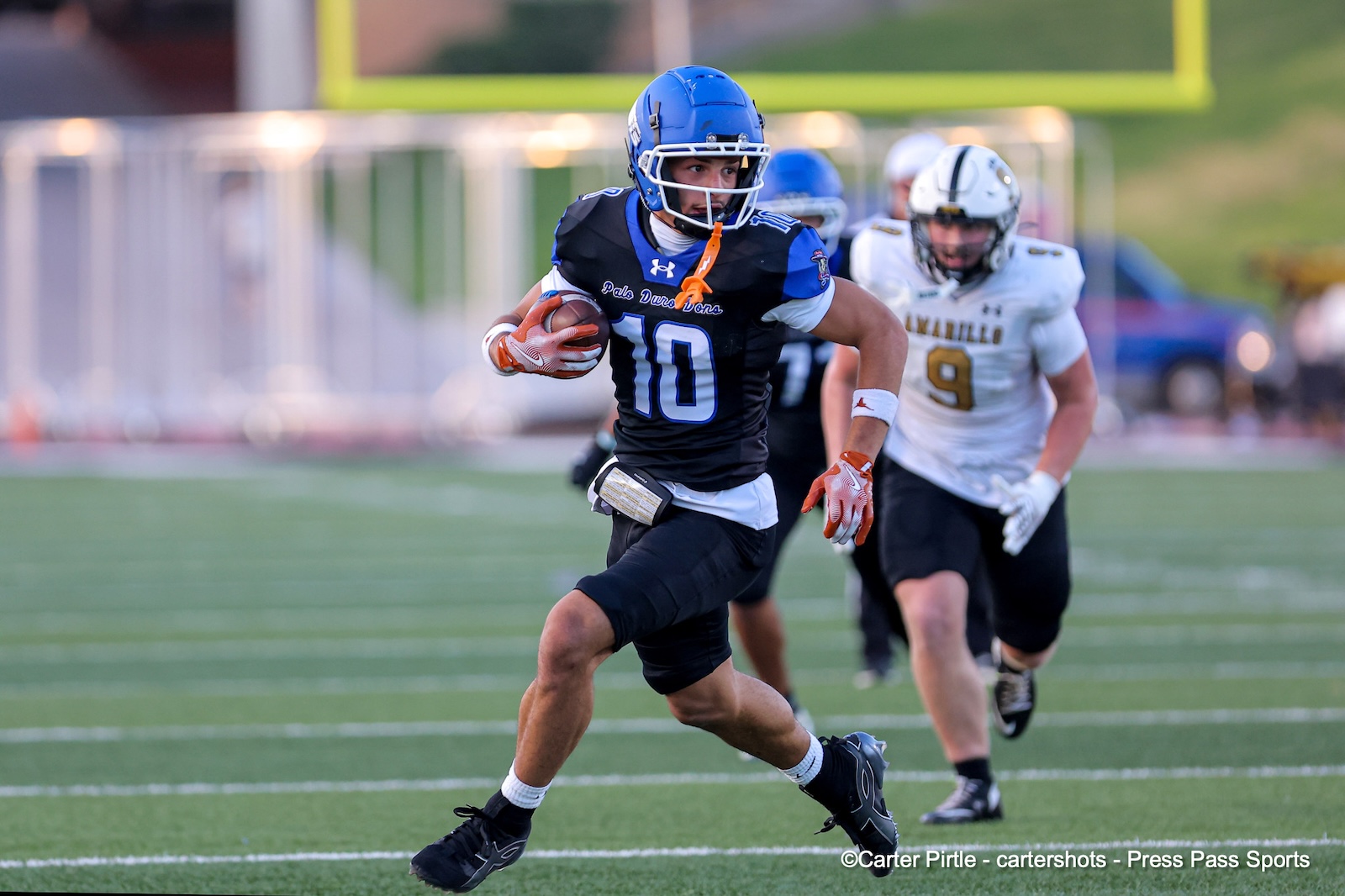 Football player in a blue helmet and black jersey with number 10 runs with the ball down the field, pursued by a white-jersey defender.