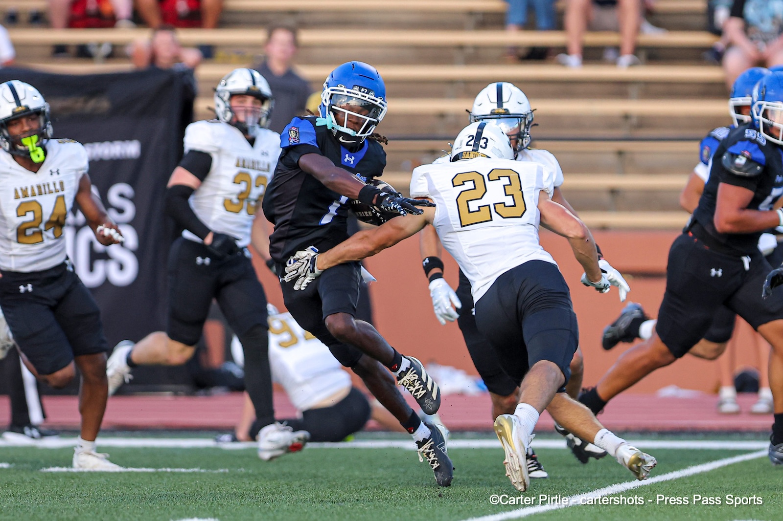 Blue-helmeted football player carries the ball as a white-jersey defender (#23) reaches to tackle during a game on a green turf field with teammates in the background.]