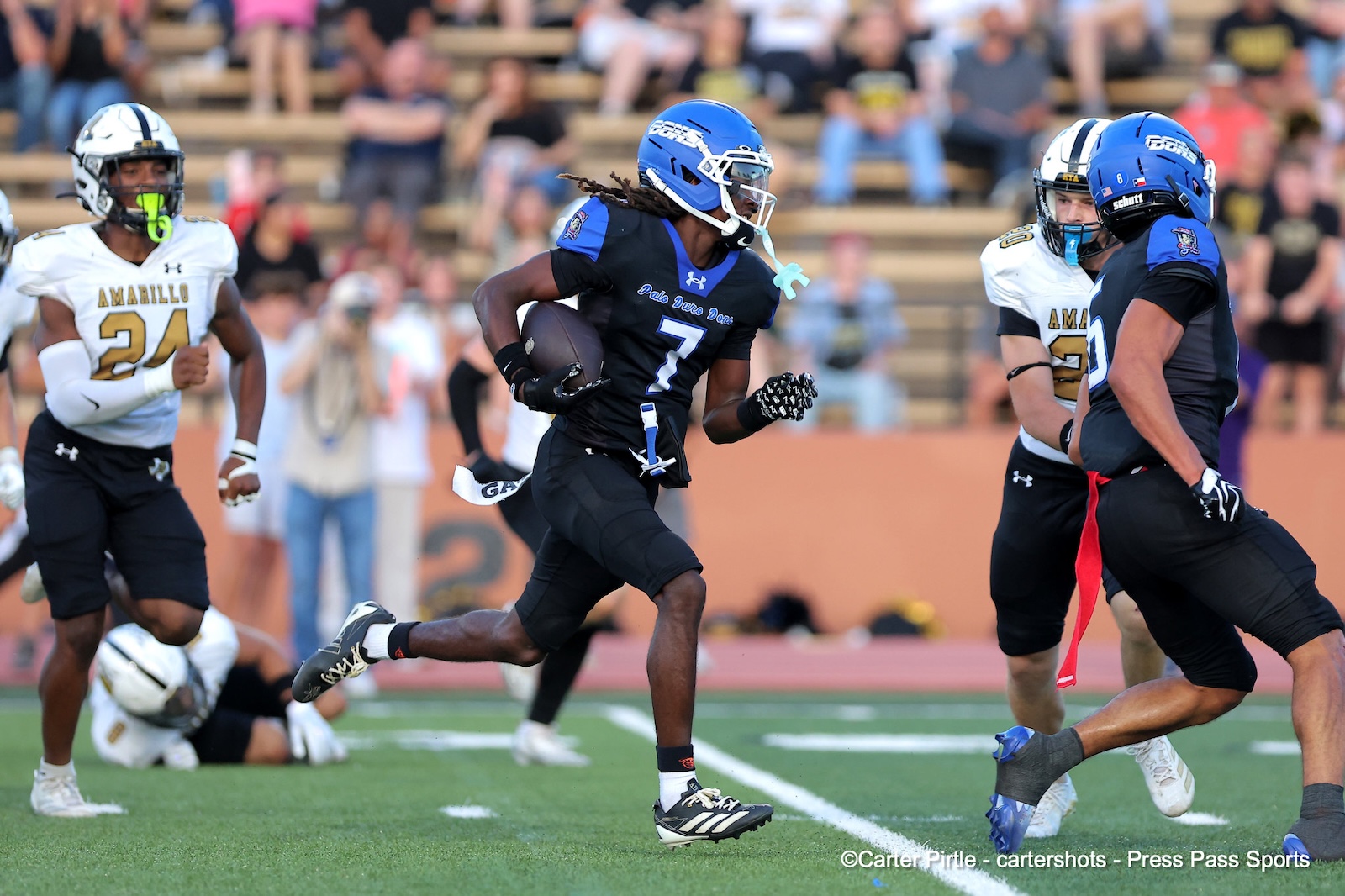 Football game action: a player wearing a blue helmet and number 7 in black pads carries the ball, chased by defenders on a green field.