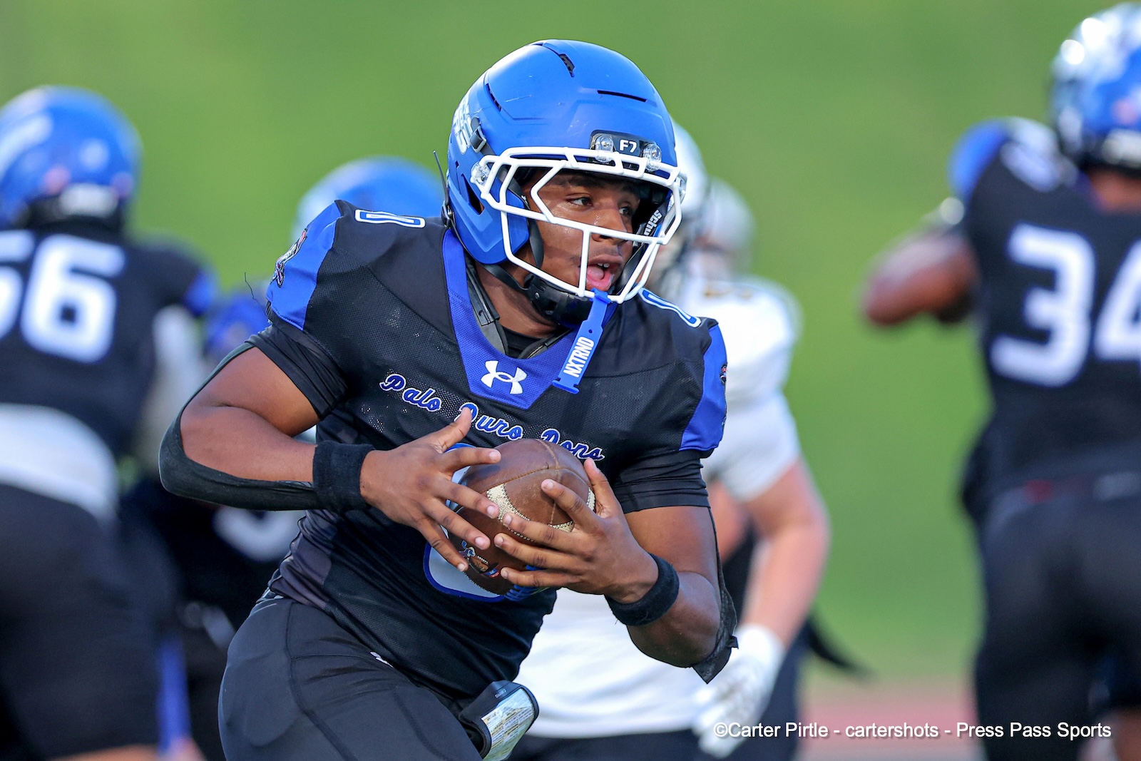 Football running back in blue helmet clutching the ball, preparing to dodge defenders on a green field.