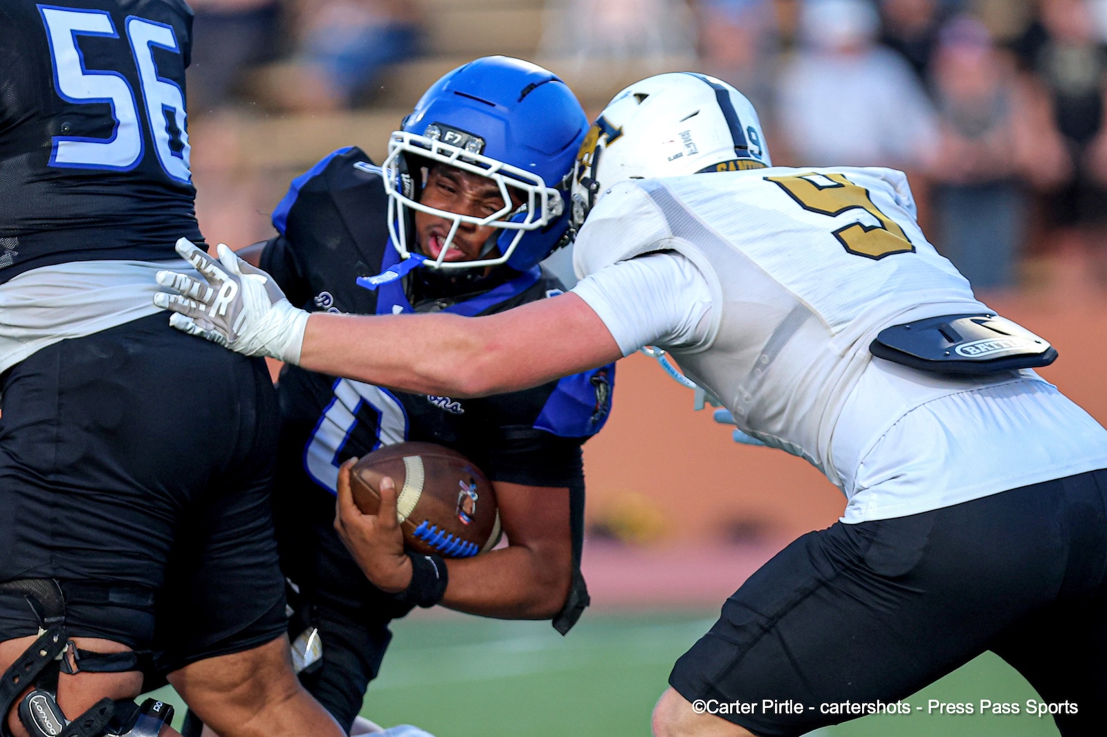 Football player in blue jersey holds the ball while being tackled by two opponents during a game.