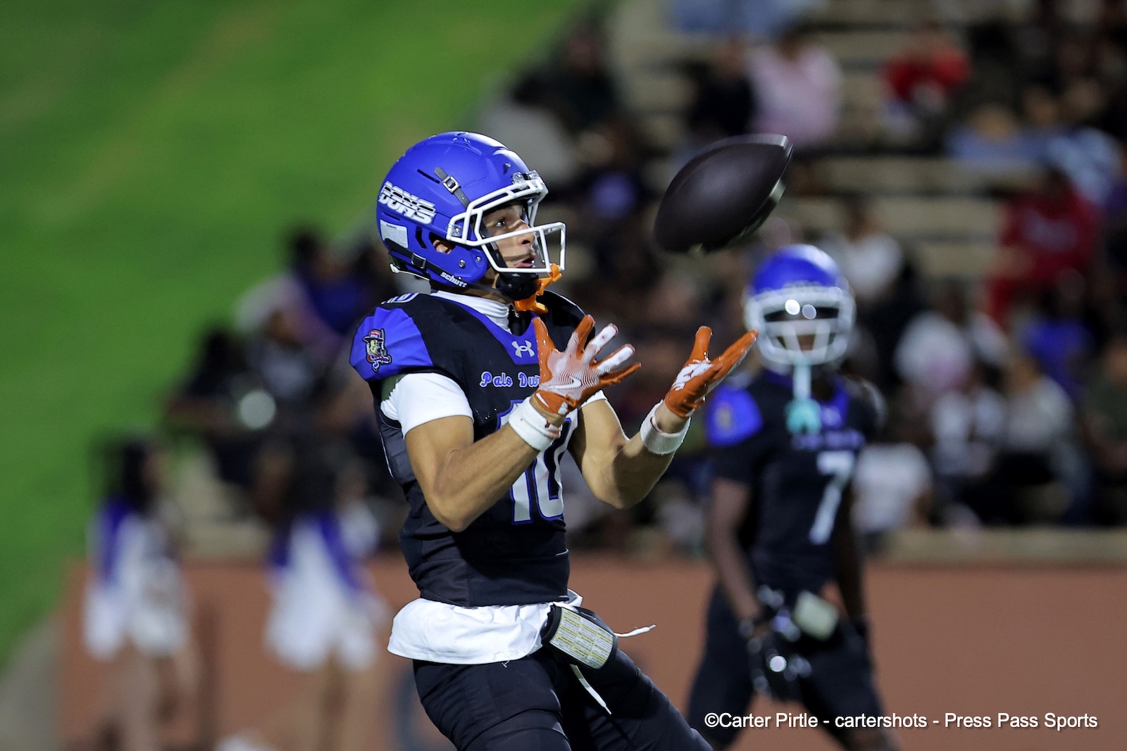 Football player in a blue helmet and dark uniform reaches for an airborne pass during a game with orange gloves visible on his hands.