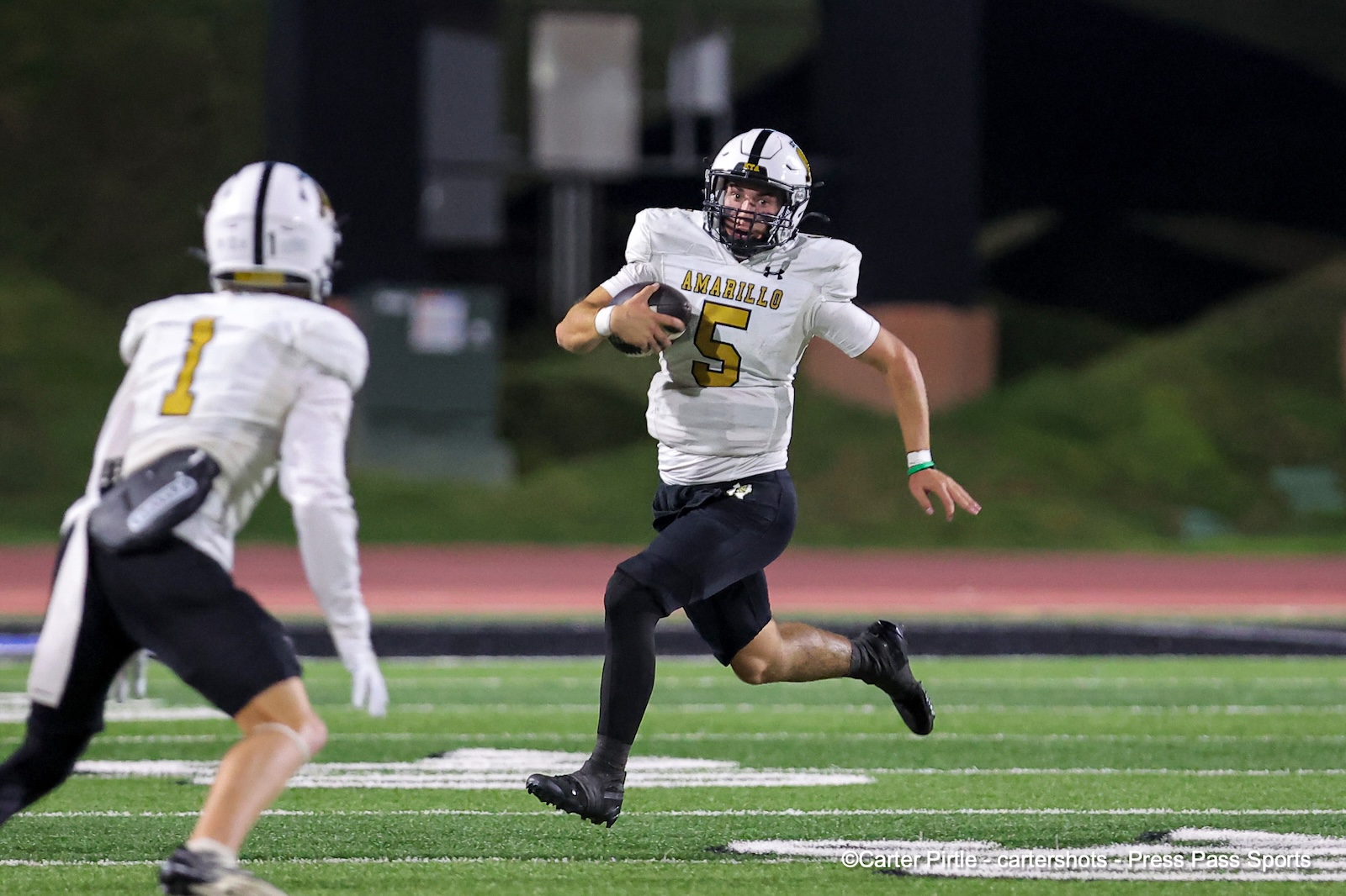 Amarillo quarterback number 5 carries the football while running past a defender on a green turf field at night.