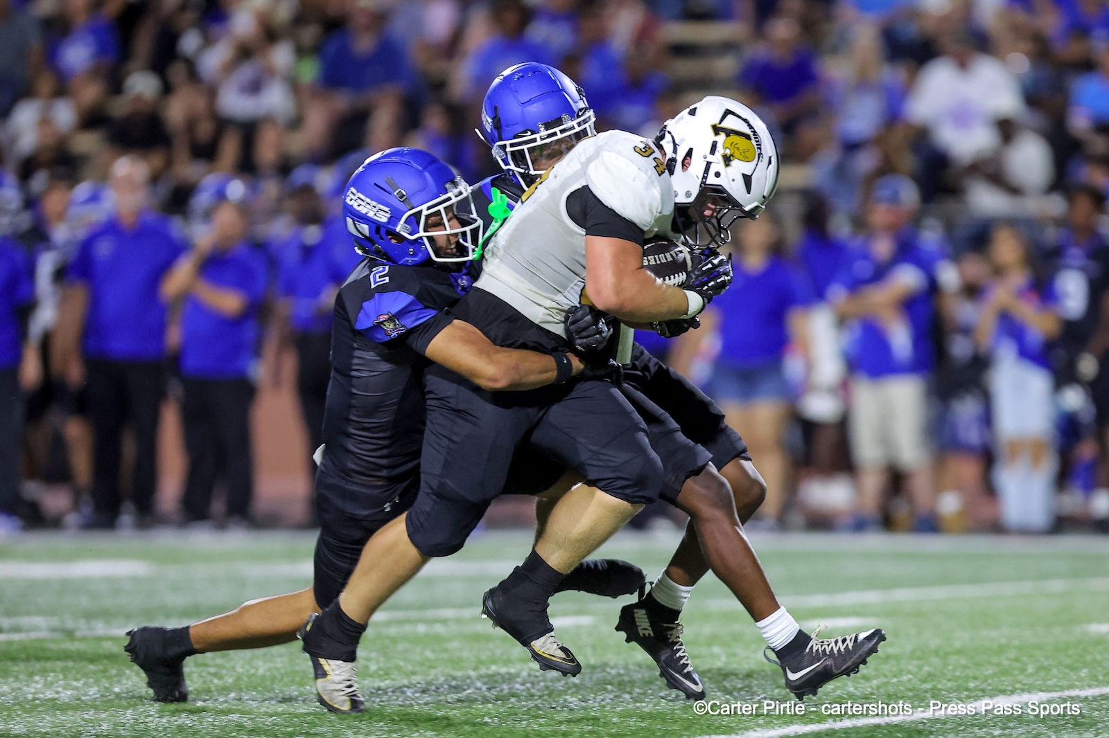 Two football players in blue uniforms tackle a white-jersey runner on a green field during a game; crowd in blue in the background.