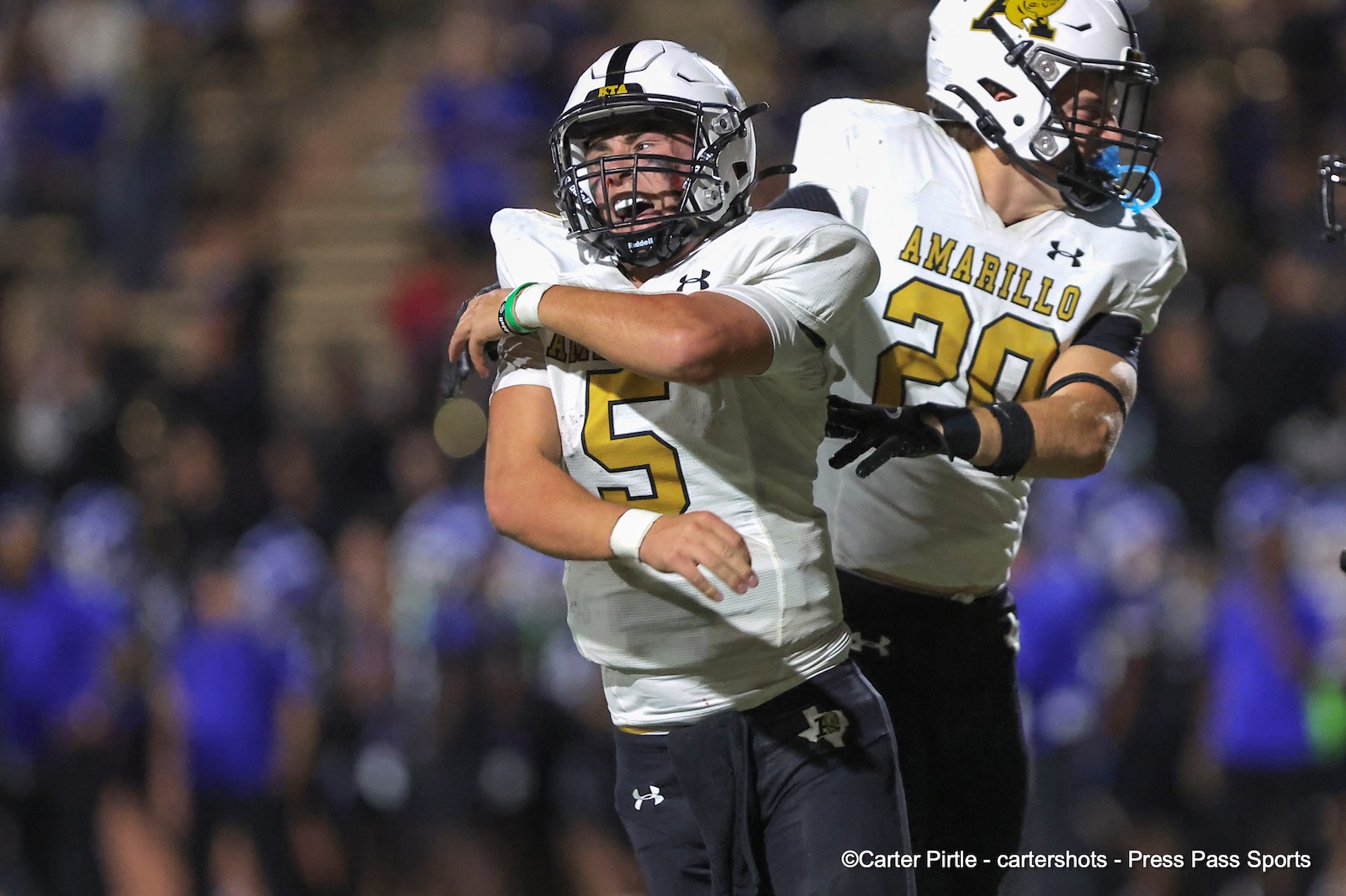 Two Amarrillo football players celebrate on the field at night, one wearing number 5 and the other number 20, in white and gold uniforms.