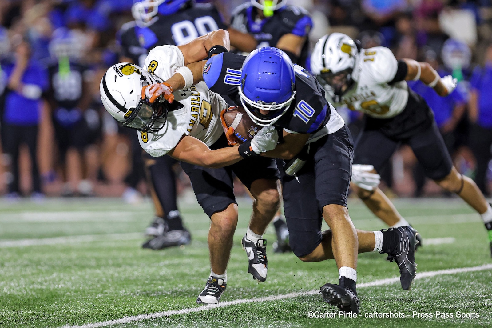 Two football players tackle each other during a game on a green turf field, with one player holding the ball in a white jersey and blue helmet nearby the clash.