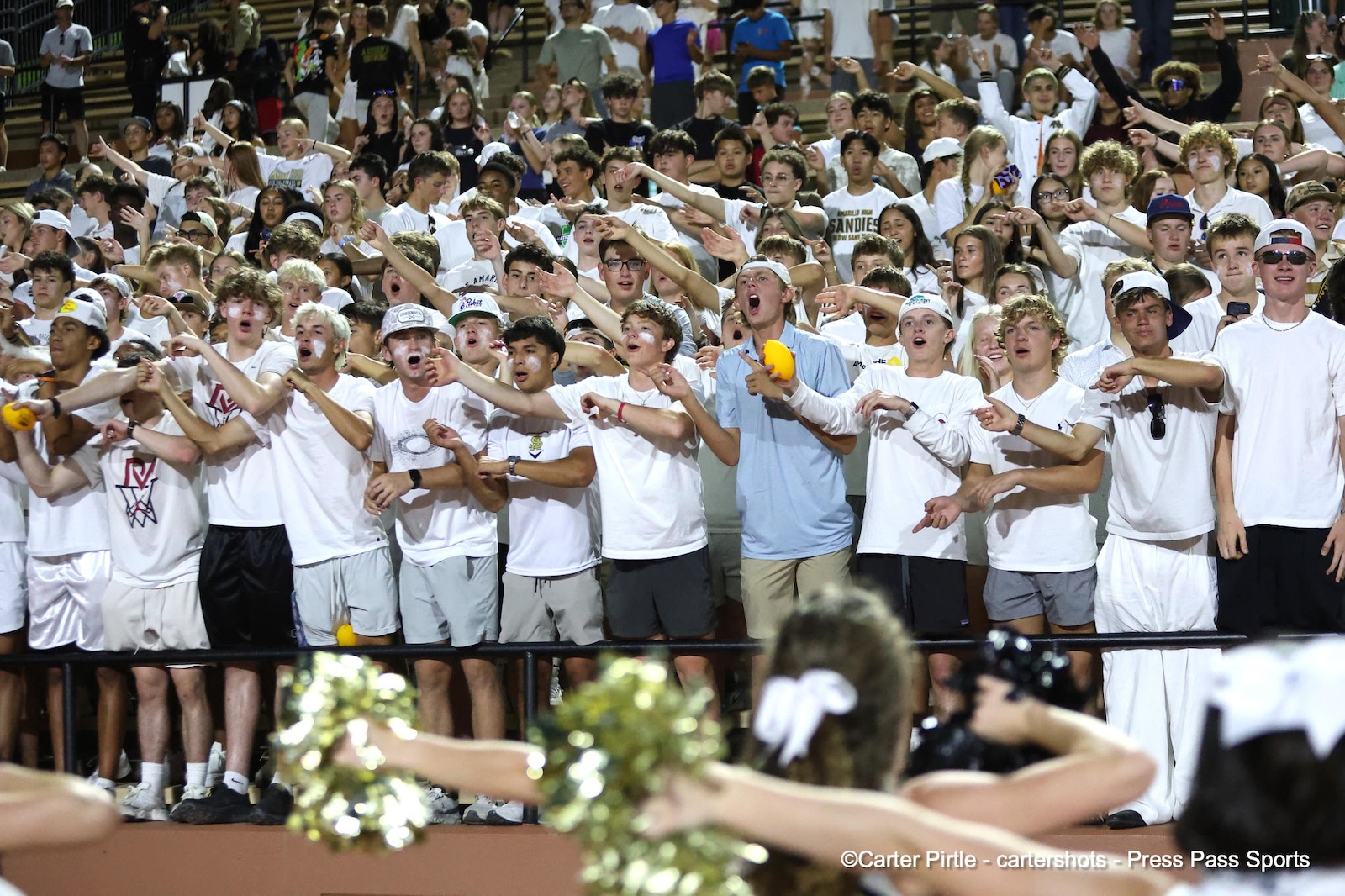 Crowd of students in white shirts cheering with arms outstretched at a stadium event, pom-poms visible in foreground.