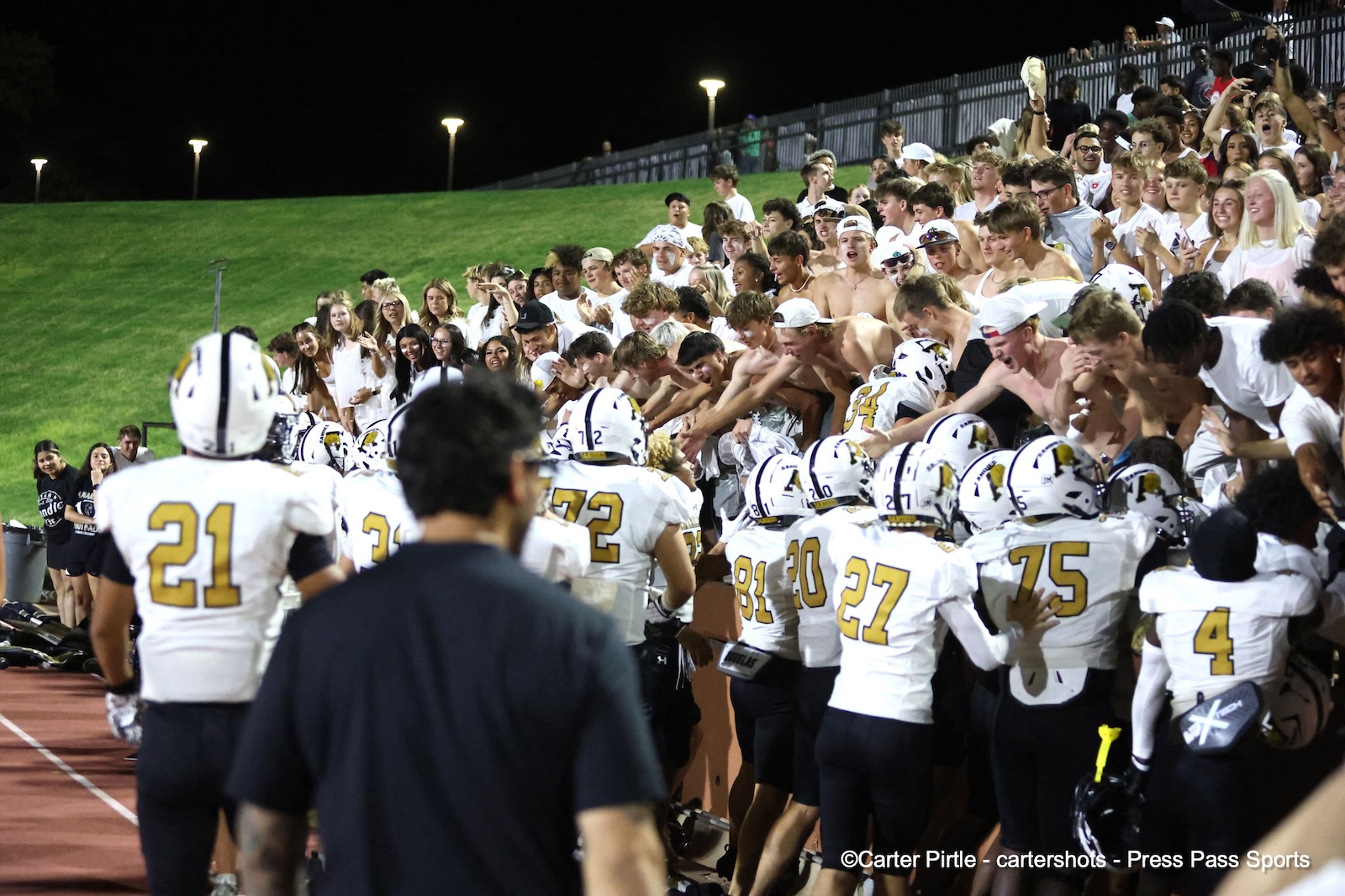 Football players in white uniforms huddle at the sideline while a cheering crowd in white watches from the bleachers at night.