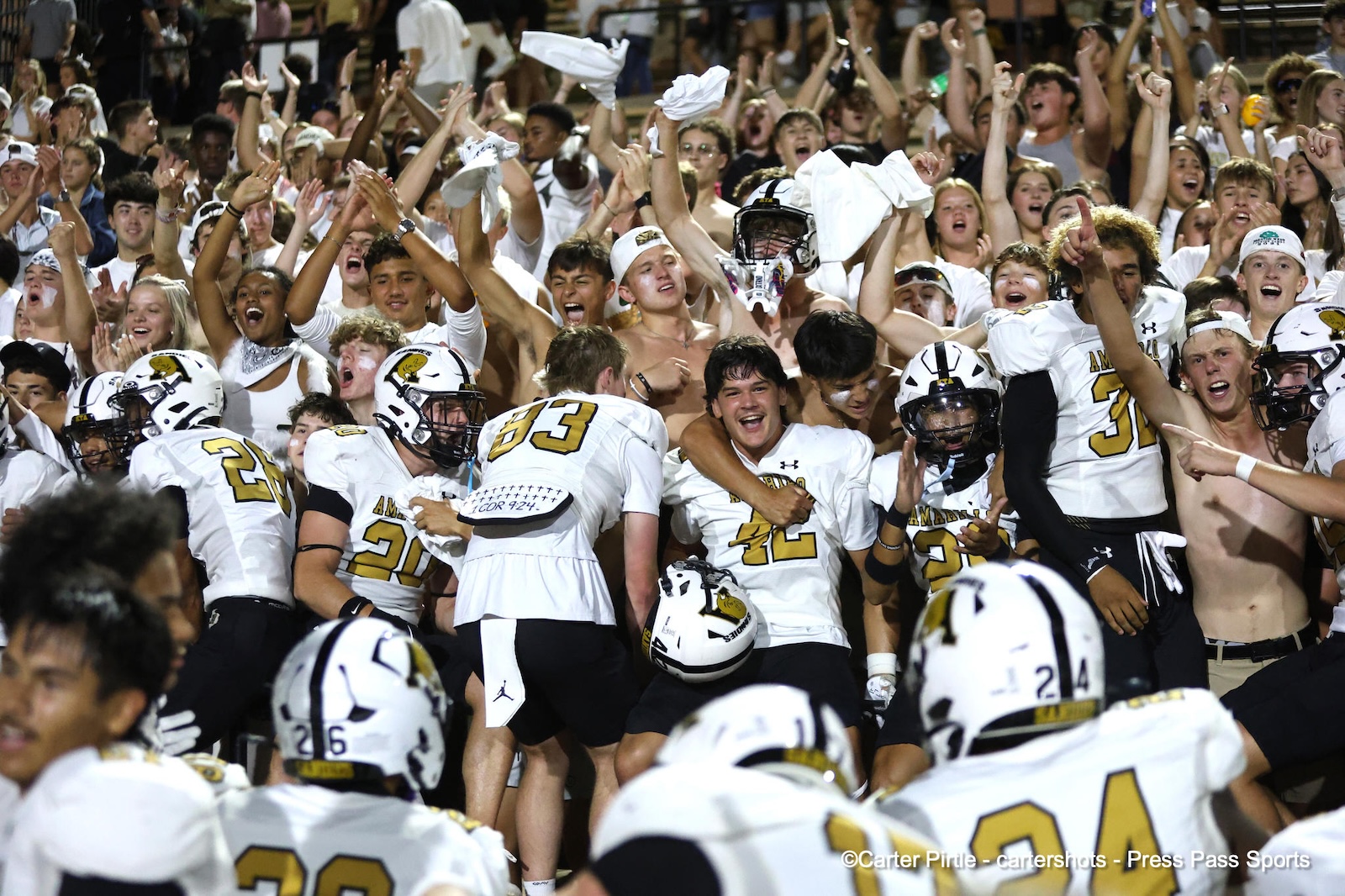 College football players in white and gold jerseys celebrate on the field while the crowd cheers with raised hands behind them, some shirtless fans visible in white shirts or face paint.