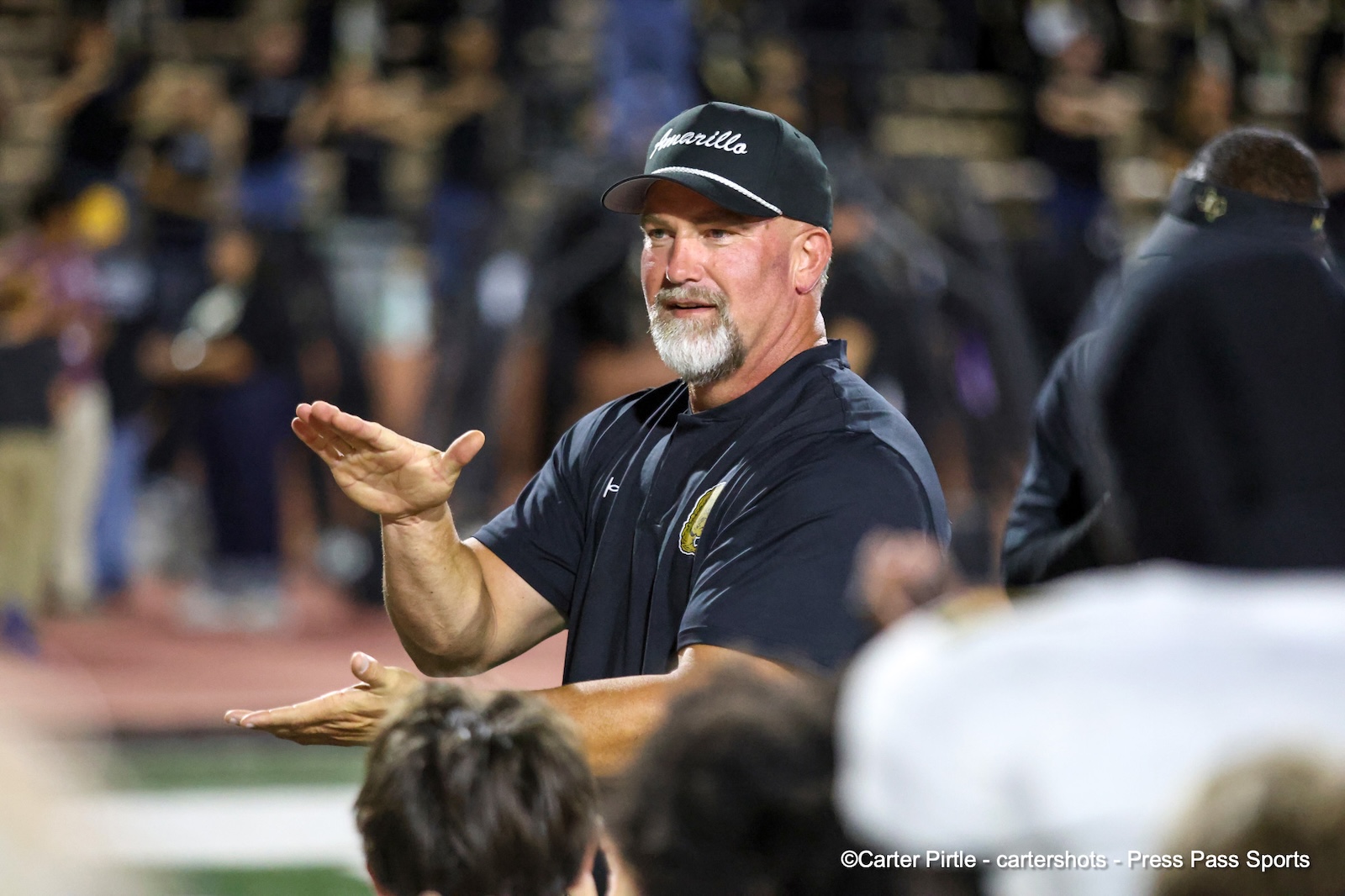 Coach on the sideline, wearing a dark shirt and cap, gesturing with his hands during a game.
