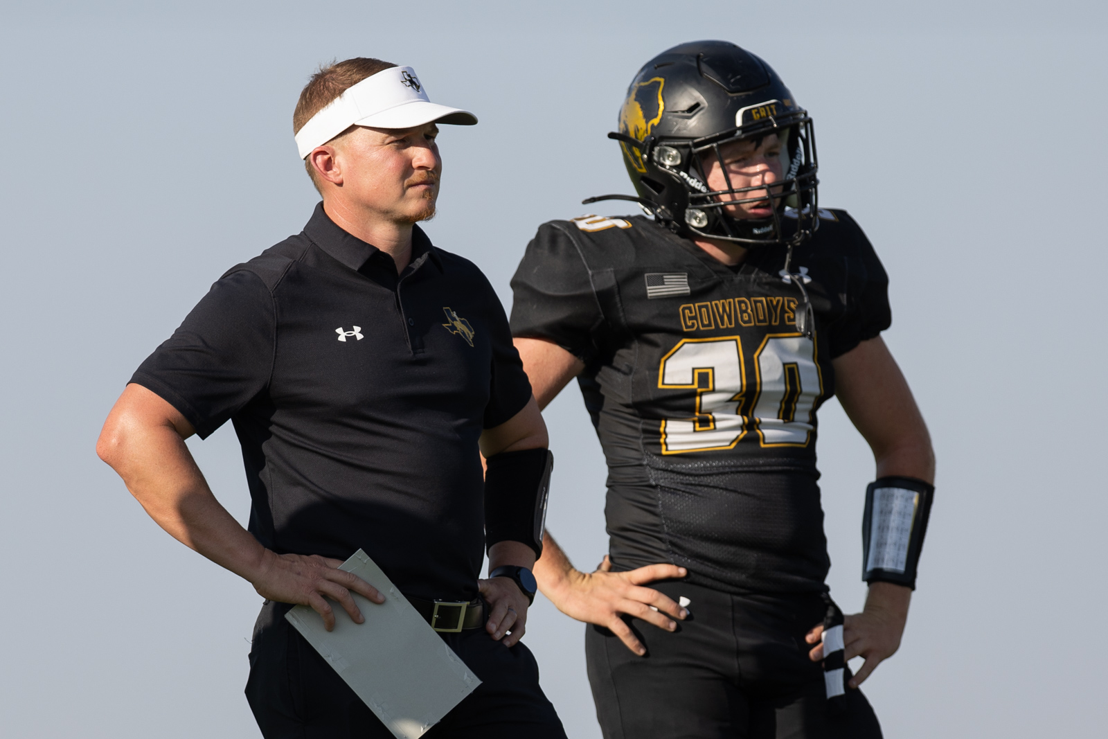 HHS-NHS-FB-2 | Press Pass Sports Football coach in black polo and white visor stands beside a player in full Cowboys football gear, clipboard in hand.