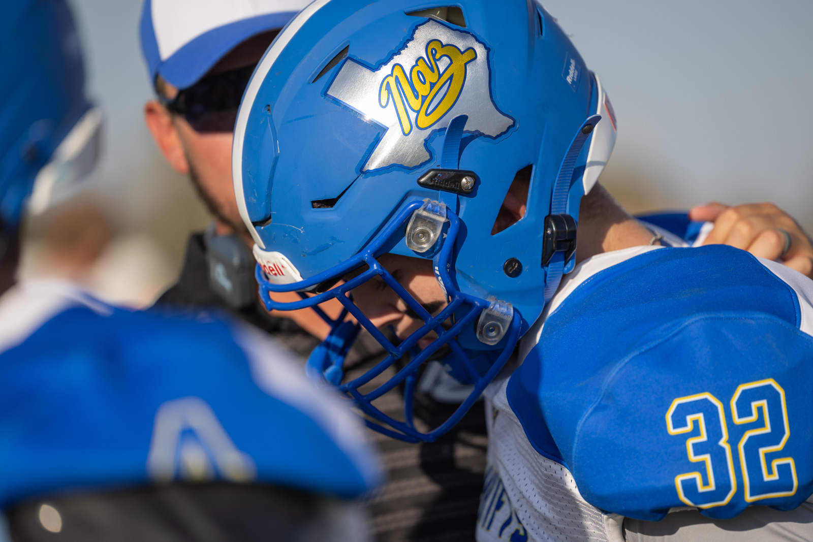 HHS-NHS-FB-3 | Press Pass Sports Close-up of a blue lacrosse helmet and shoulder of a player wearing number 32, as teammates hug in a blue-and-white uniform.