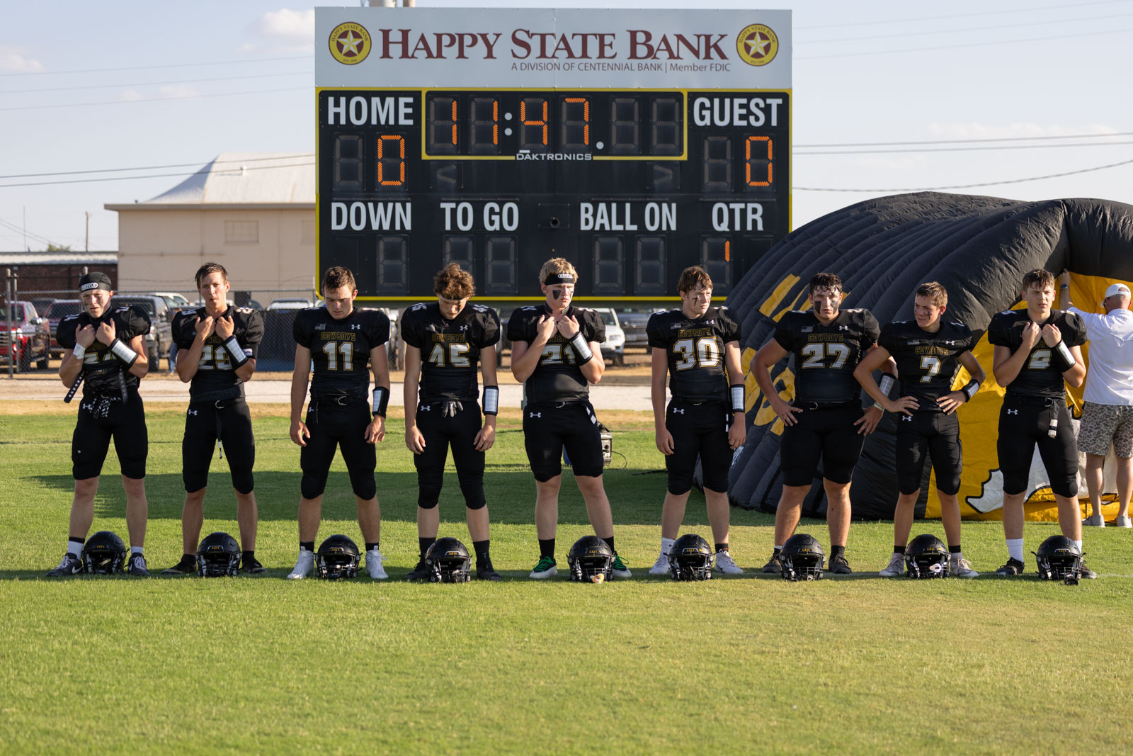 HHS-NHS-FB-5 | Press Pass Sports Line of football players in black uniforms standing on the field with helmets on the grass in front, scoreboard behind them at a sunny stadium edge.
