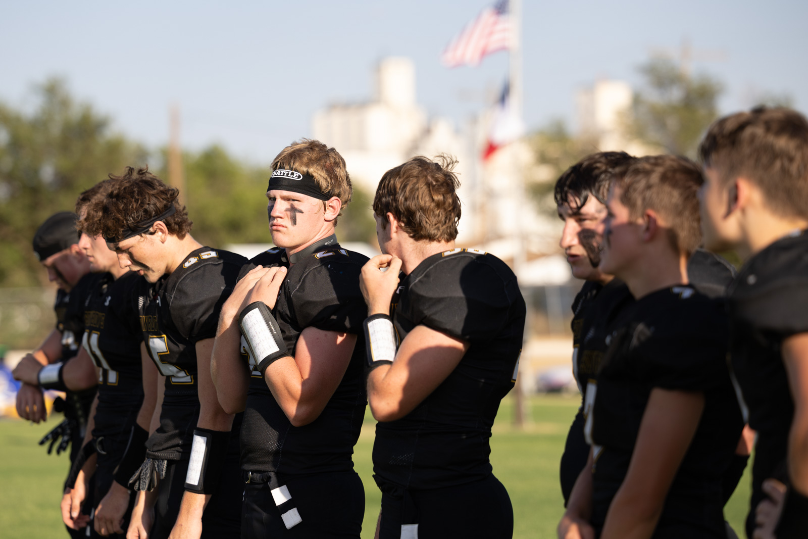 HHS-NHS-FB-6 | Press Pass Sports Line of American football players in black uniforms standing in a row on a field during warm‑ups, facing left.