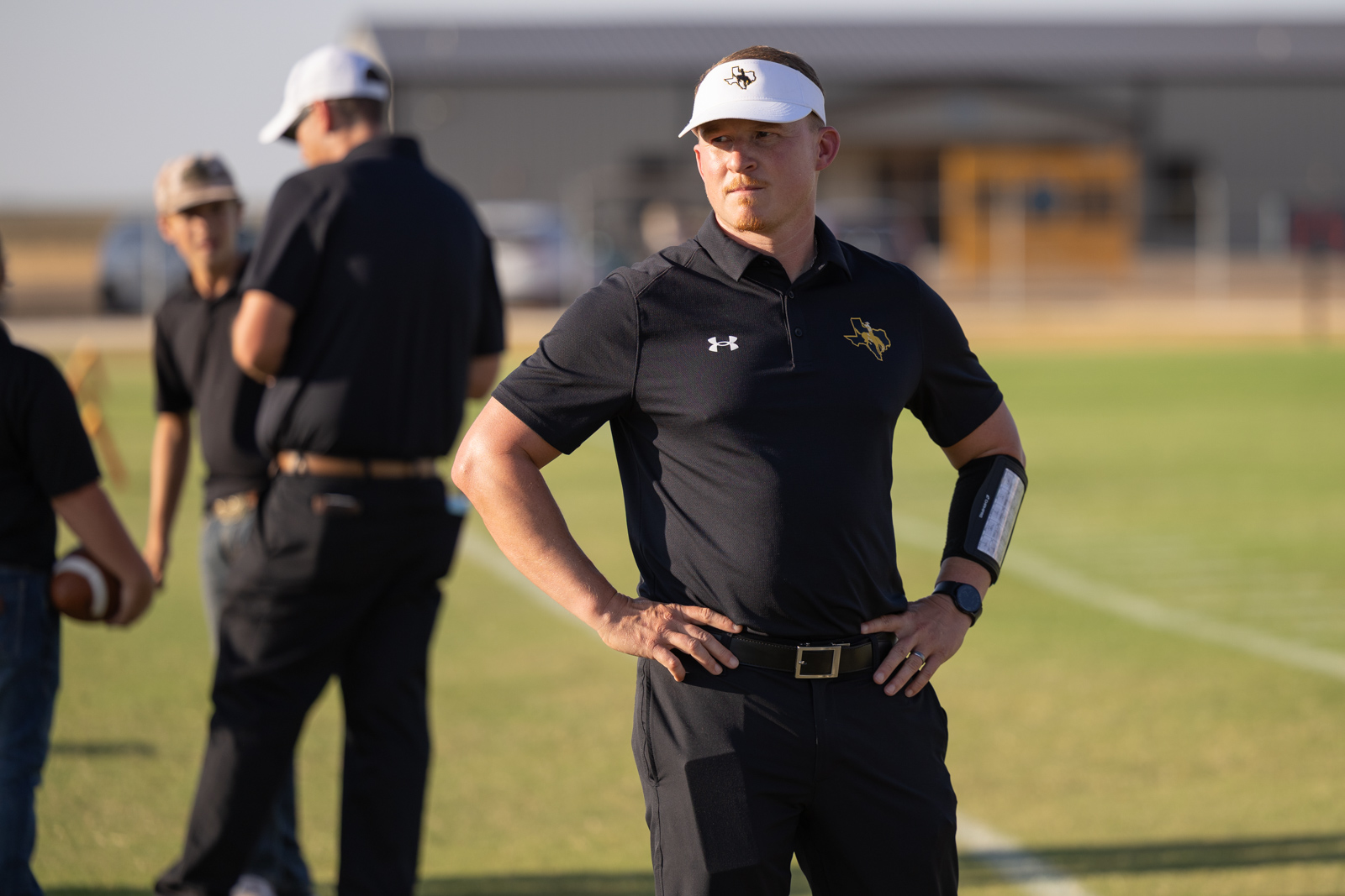 HHS-NHS-FB-7 | Press Pass Sports Coach in a black polo and white visor stands with hands on hips on a football field.