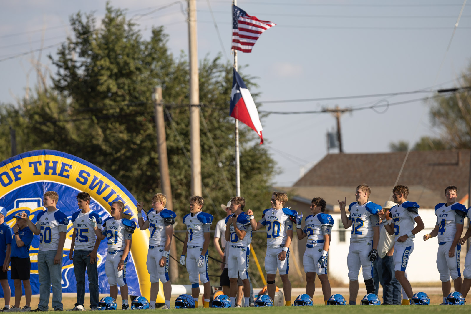 HHS-NHS-FB-8 | Press Pass Sports Line of football players in white and blue uniforms standing on the sideline with helmets on the grass and a large blue-yellow arch behind them.