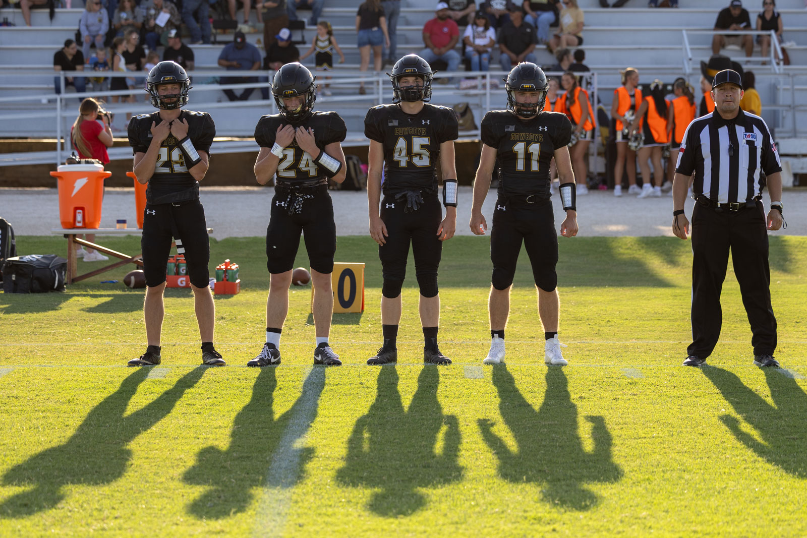 HHS-NHS-FB-9 | Press Pass Sports Five football players in black uniforms stand in a line on a grassy field while a referee watches, with spectators in the bleachers behind them.