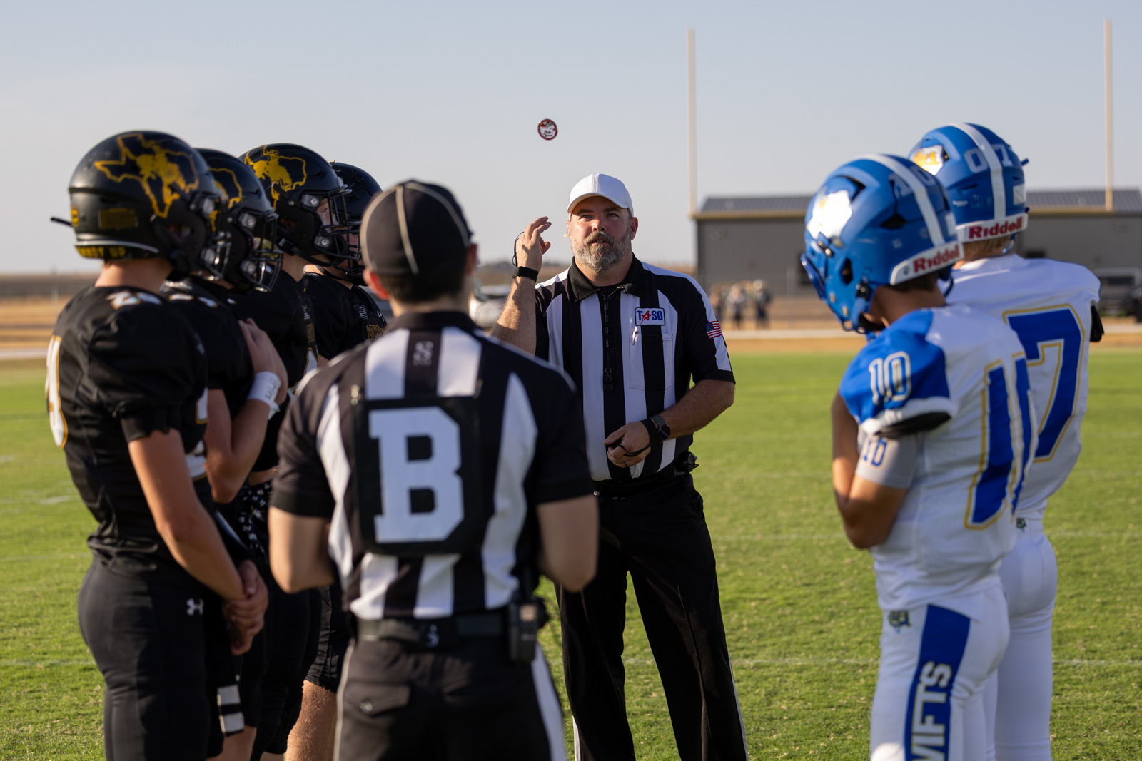 HHS-NHS-FB-12 | Press Pass Sports Referee performs a coin toss between black-uniformed team captains (left) and white-blue team captains (right) on a football field.