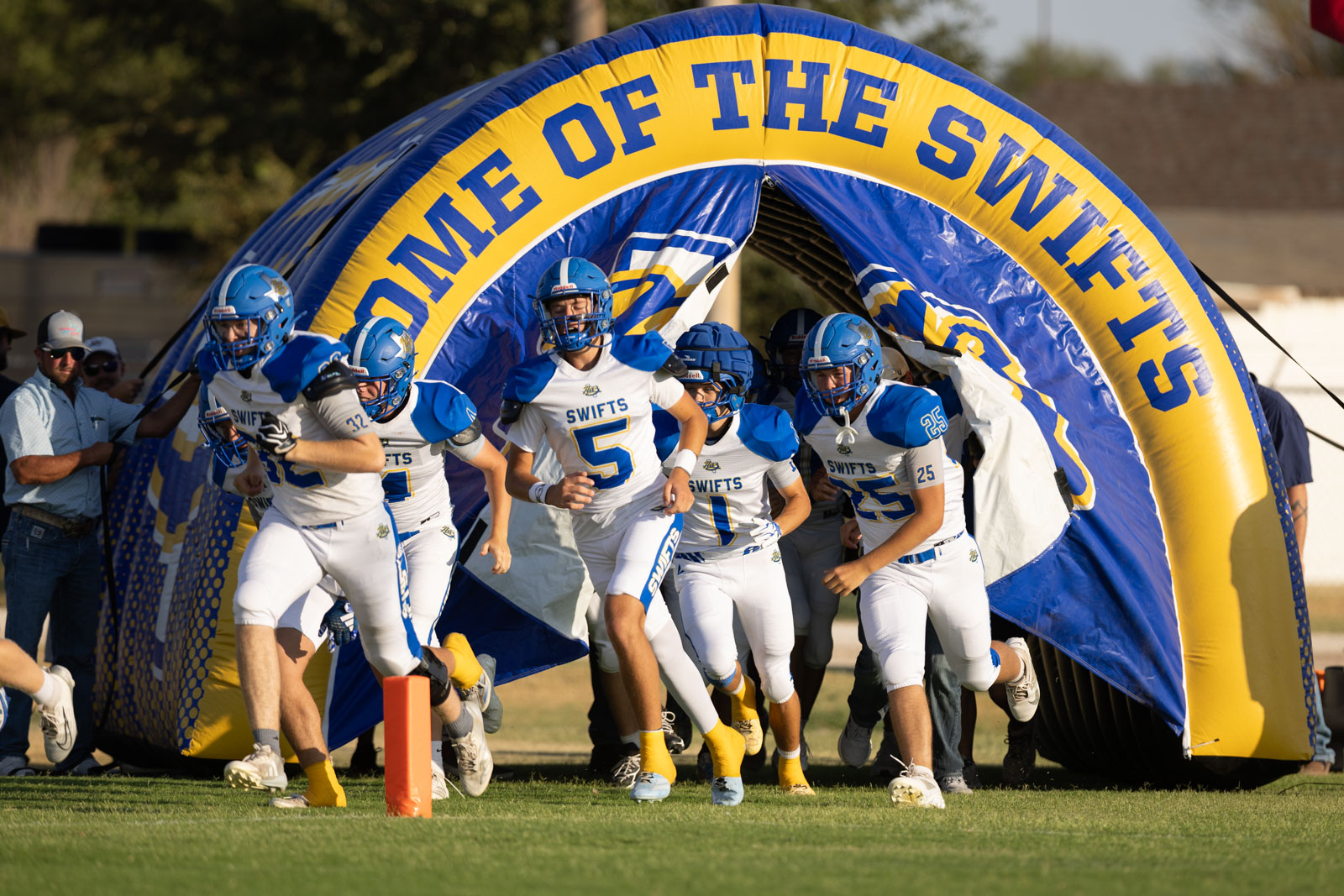 HHS-NHS-FB-15 | Press Pass Sports Youth football players in white and blue uniforms sprint onto the field through a blue-and-yellow inflatable tunnel labeled SwiftS/Swift team, surrounded by coaches and spectators.