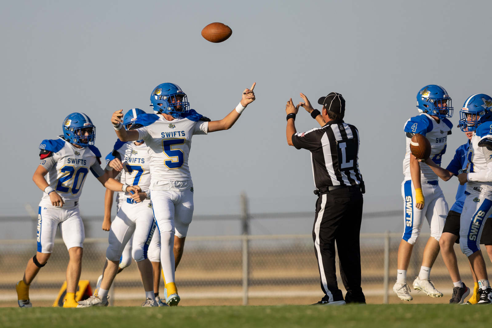 HHS-NHS-FB-17 | Press Pass Sports Football players in white and blue uniforms on a sunny field, the ball in midair as a player reaches upward and a referee signals nearby.