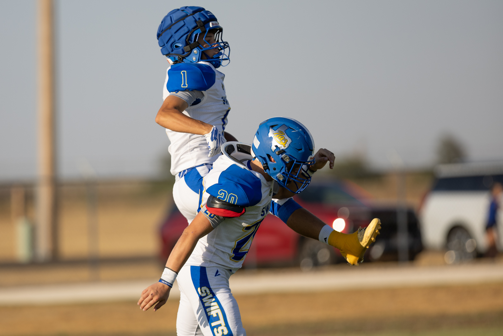 HHS-NHS-FB-19 | Press Pass Sports Two football players in blue and white uniforms collide on a sunny field; one is airborne during a tackle or block.