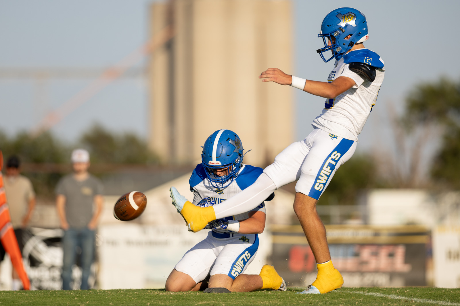HHS-NHS-FB-20 | Press Pass Sports Two Swift teammates in blue helmets and white uniforms collide on a grassy football field as the ball flies between them.