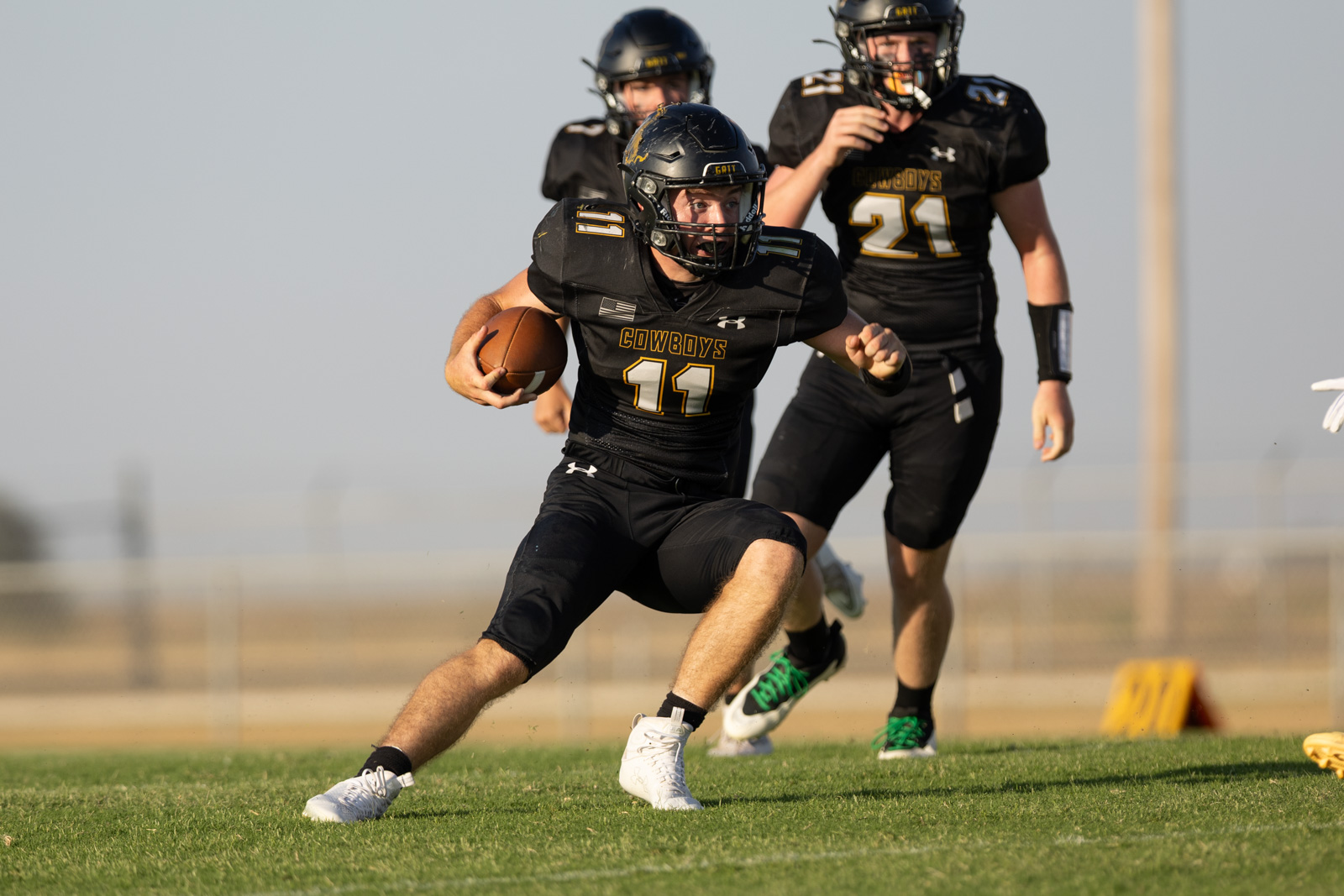 HHS-NHS-FB-22 | Press Pass Sports Football player in black Cowboys uniform with number 11 runs with the ball on a grassy field, teammates in pursuit behind him