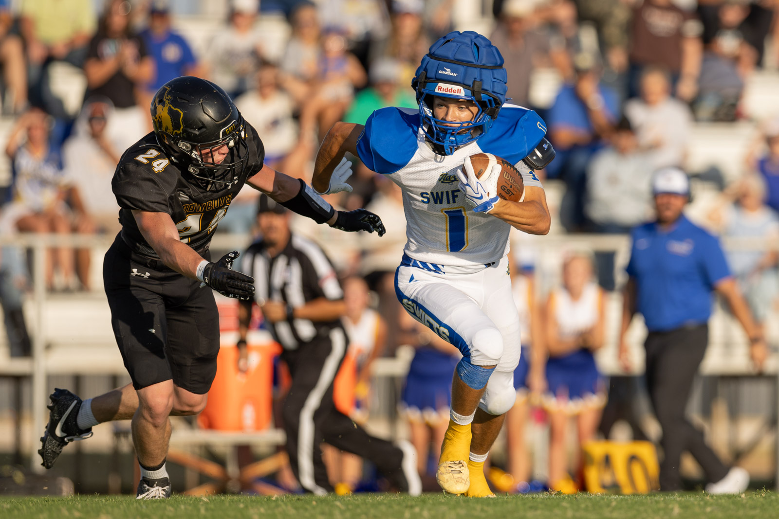 HHS-NHS-FB-24 | Press Pass Sports Football player in white/blue jersey runs with the ball while a defender in black chases behind, with a crowded stands background.