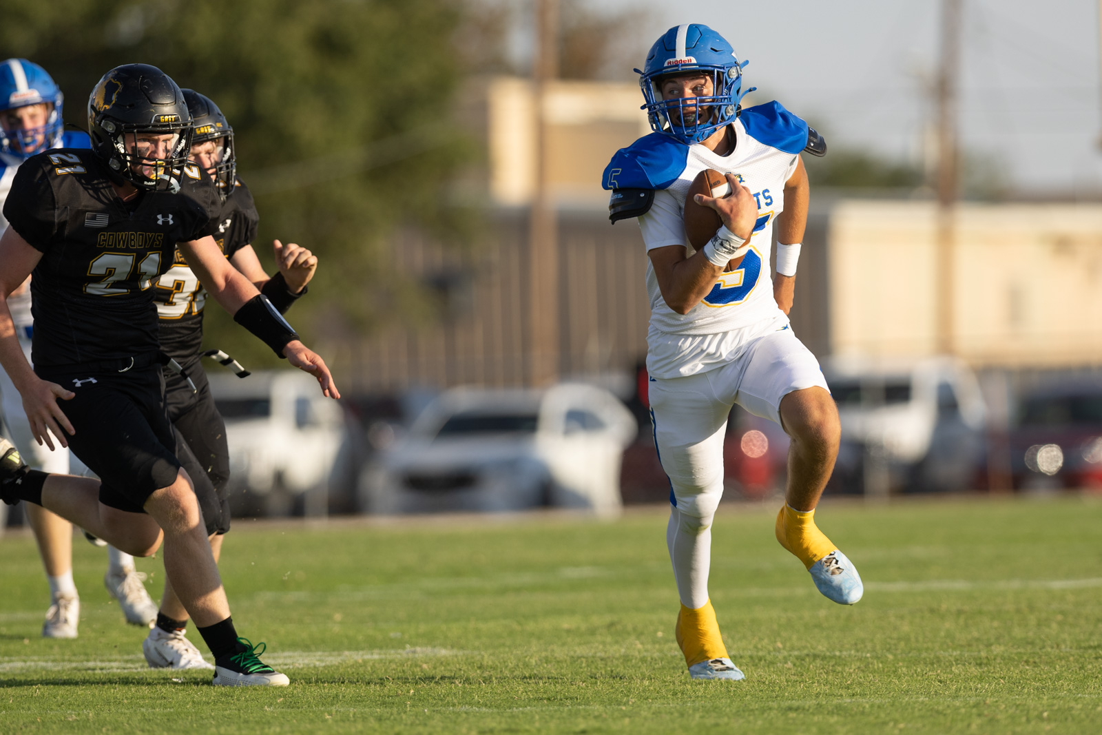 HHS-NHS-FB-25 | Press Pass Sports Football game action: a ball-carrier in a white and blue uniform runs with the ball as black-clad defenders chase him down the field.