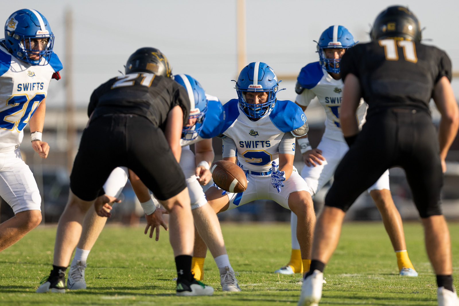HHS-NHS-FB-26 | Press Pass Sports Swift #3, in blue and white, holds the football and crouches for a run as teammates and defenders close in on a sunny field