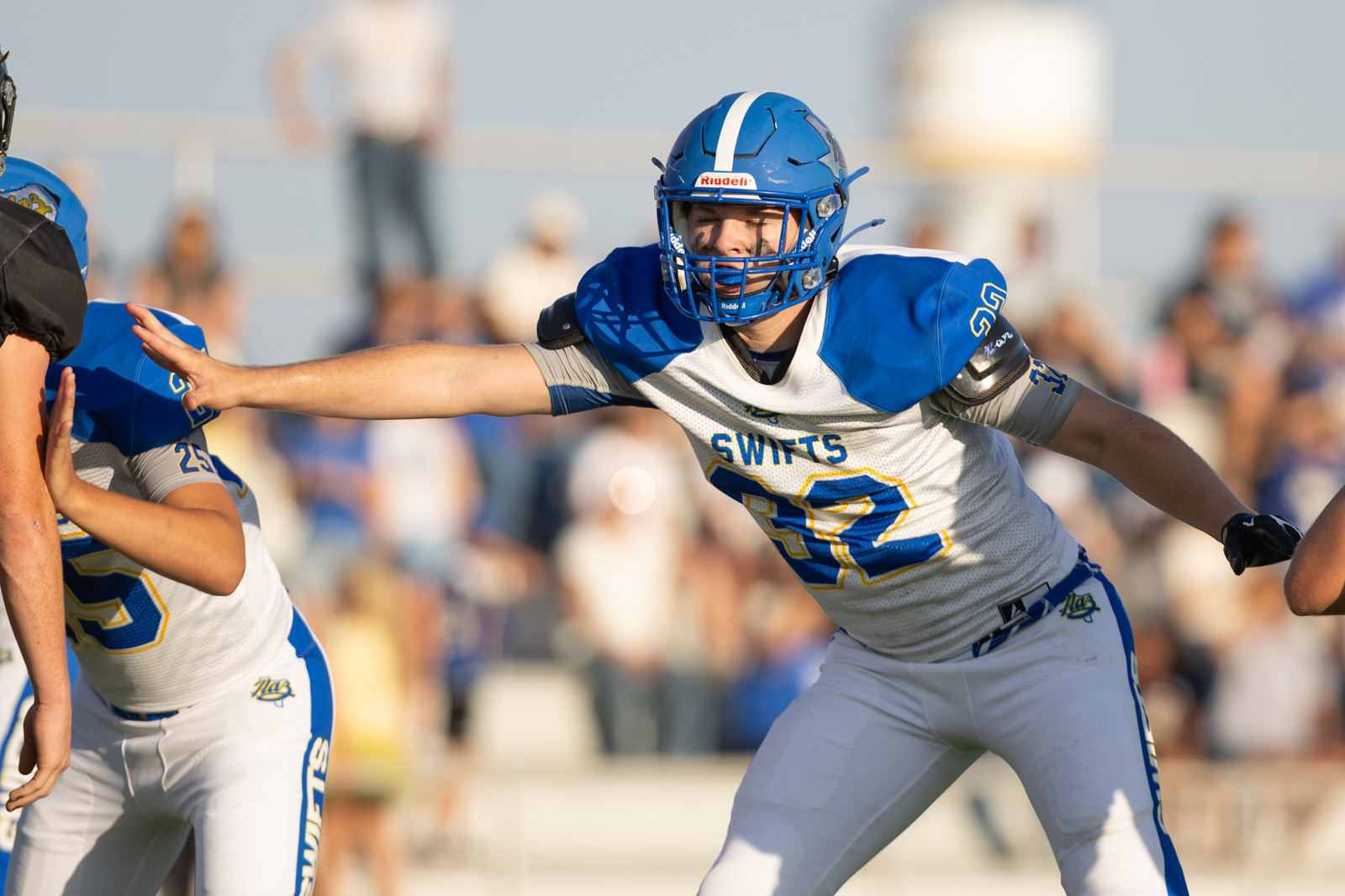HHS-NHS-FB-27 | Press Pass Sports Football players in blue and white uniforms clash at the line of scrimmage as a Swift player extends an arm to block the opponent.